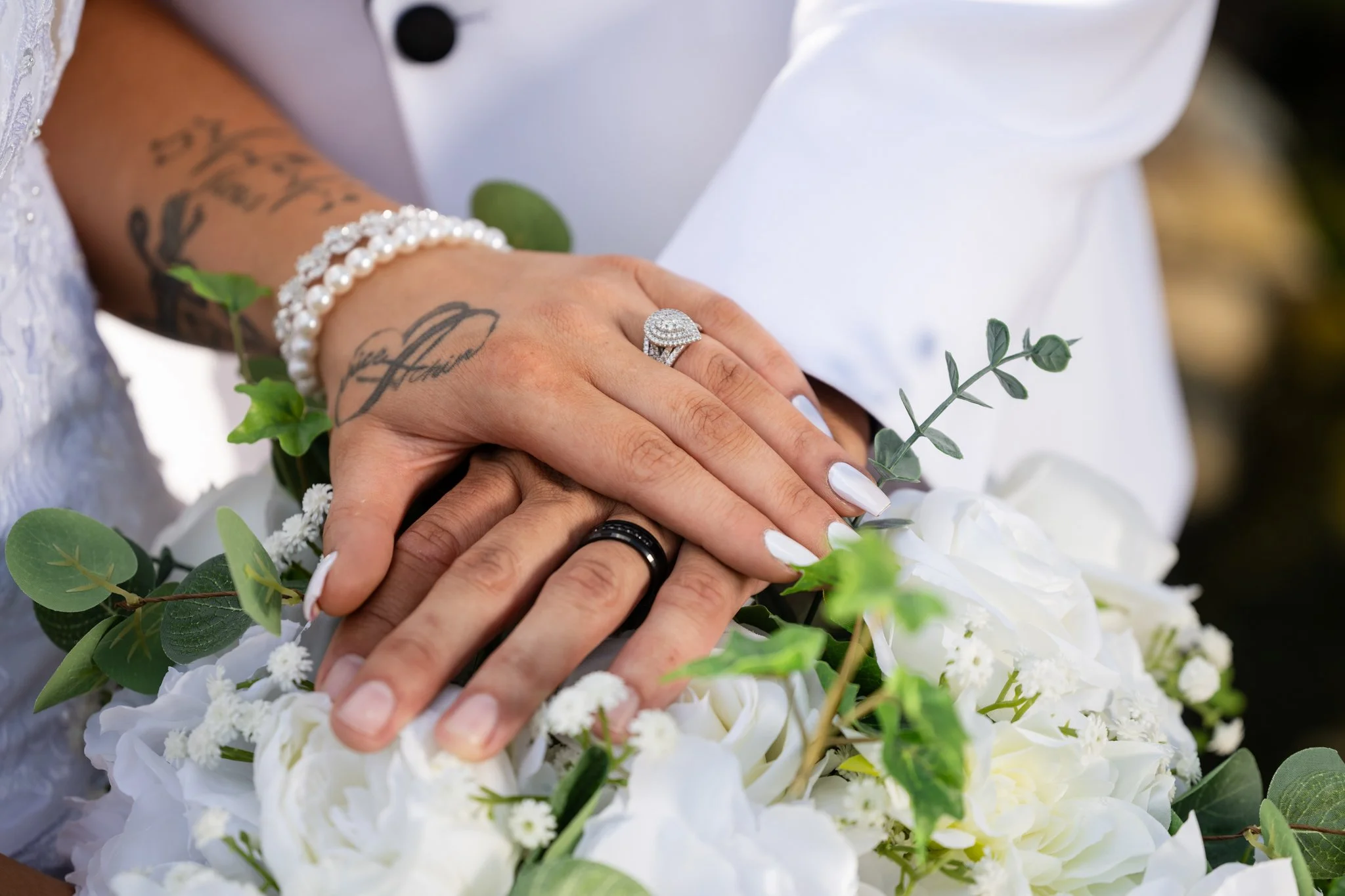 Close-up of a bride and groom's hands resting on a bouquet of white flowers and greenery, showing wedding rings and jewelry.