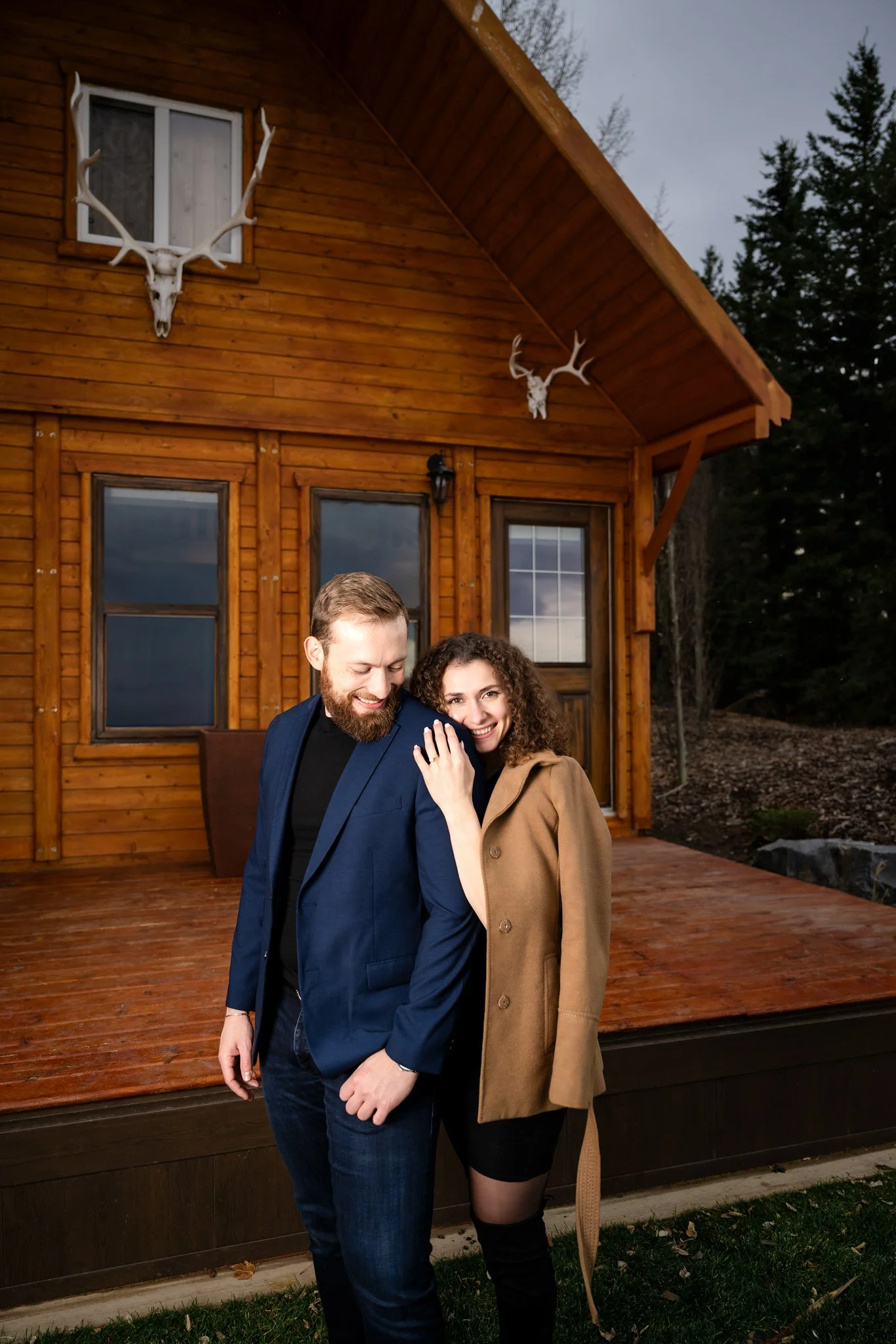 A smiling woman with curly hair showing her engagement ring, leaning on a man with a beard, both dressed in stylish clothes, standing on a wooden house porch with deer skulls and antlers mounted on the house wall behind them.