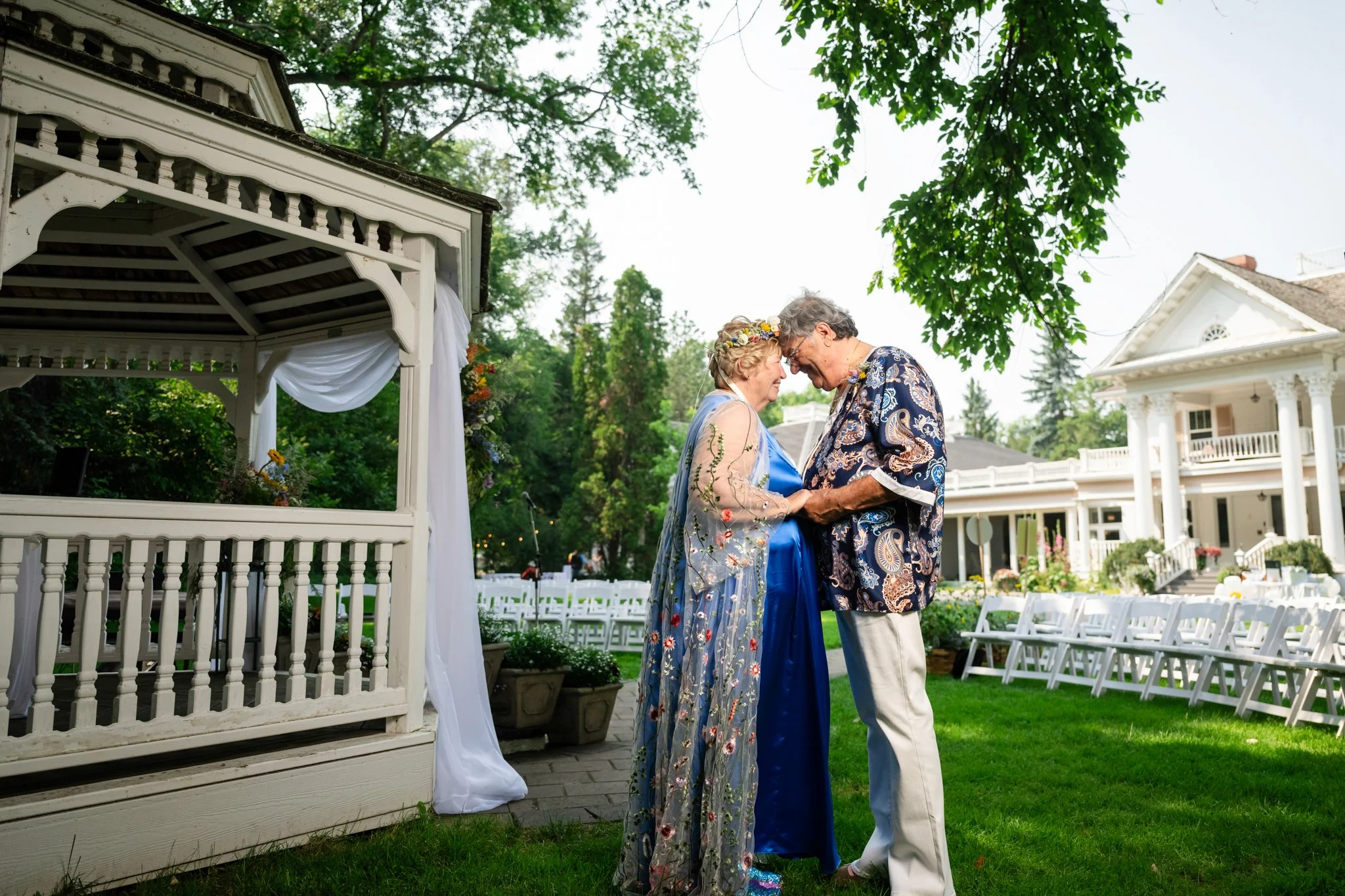 An elderly couple with glasses, dressed in wedding attire, sharing a tender moment outdoors during a wedding celebration at a white mansion with chairs and greenery in the background.