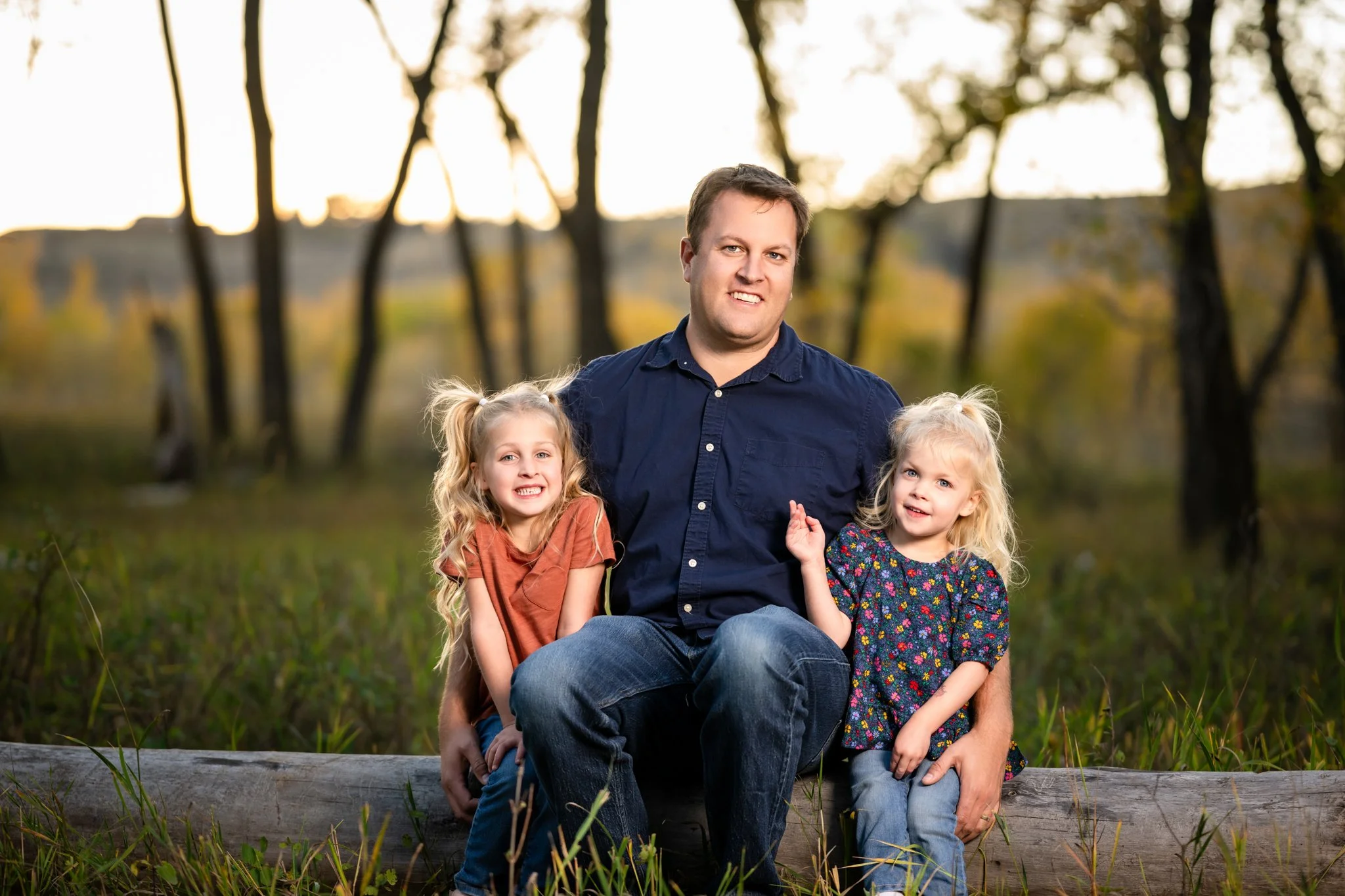 A man with two young girls sitting on a log outdoors during sunset in a wooded area.
