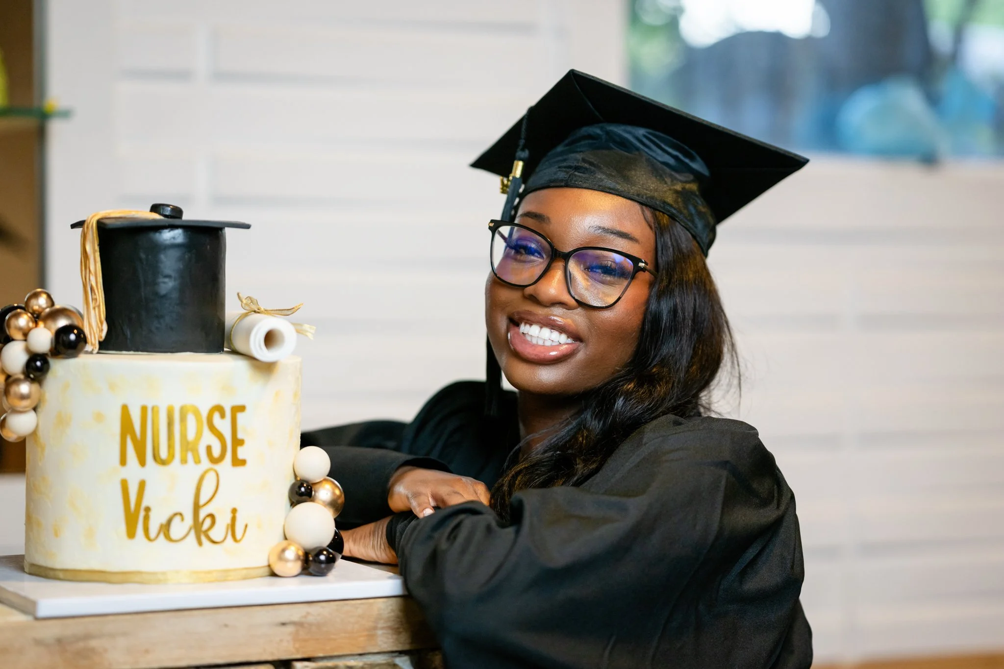 A woman in a graduation cap and gown smiling next to a decorated cake with the words "Nurse Vicki" on it.