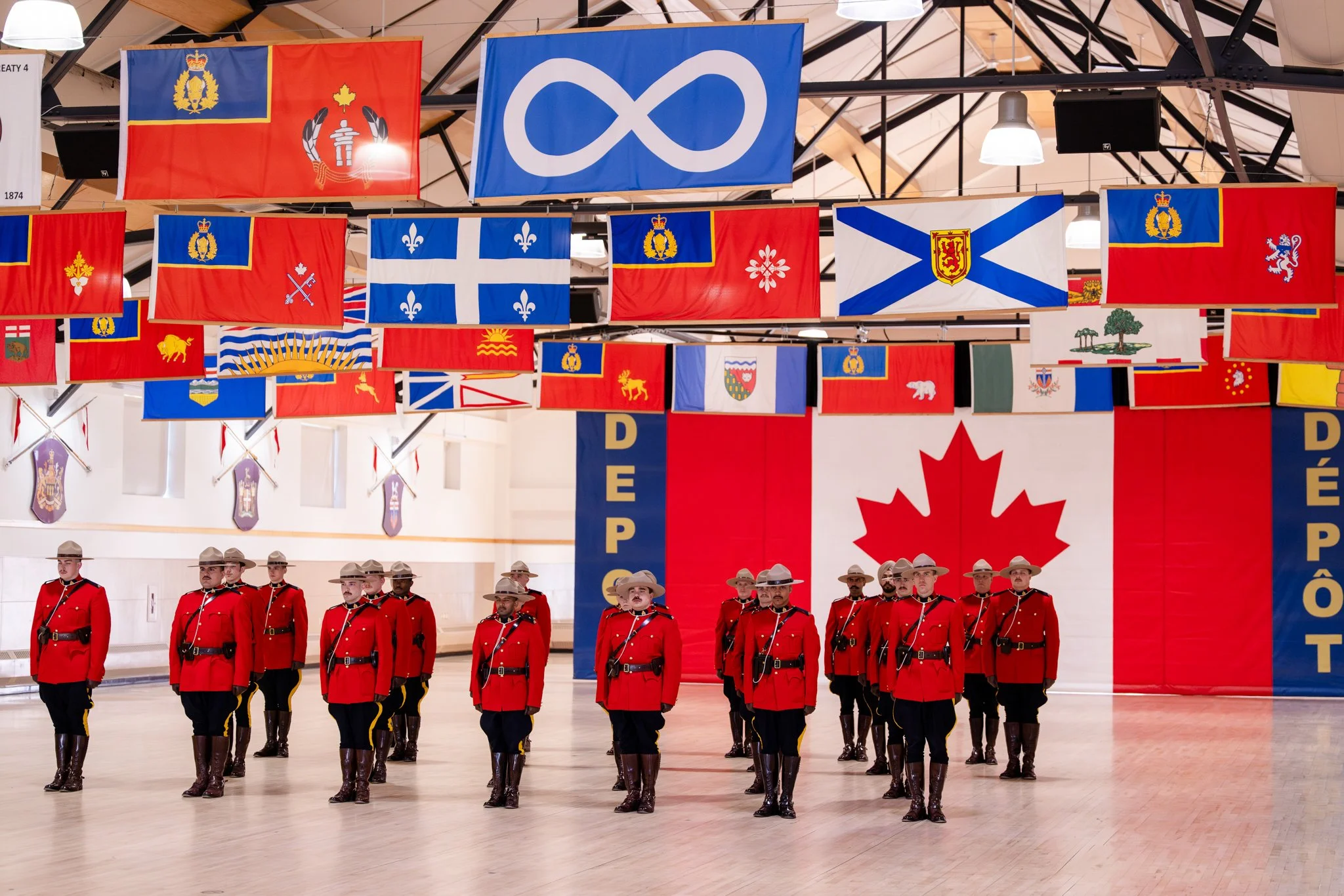 Royal Canadian Mounted Police officers in red uniforms and hats conducting a drill inside a hall decorated with various flags of different countries, with a large Canadian flag and a banner reading 'DΓPΓT' in the background.