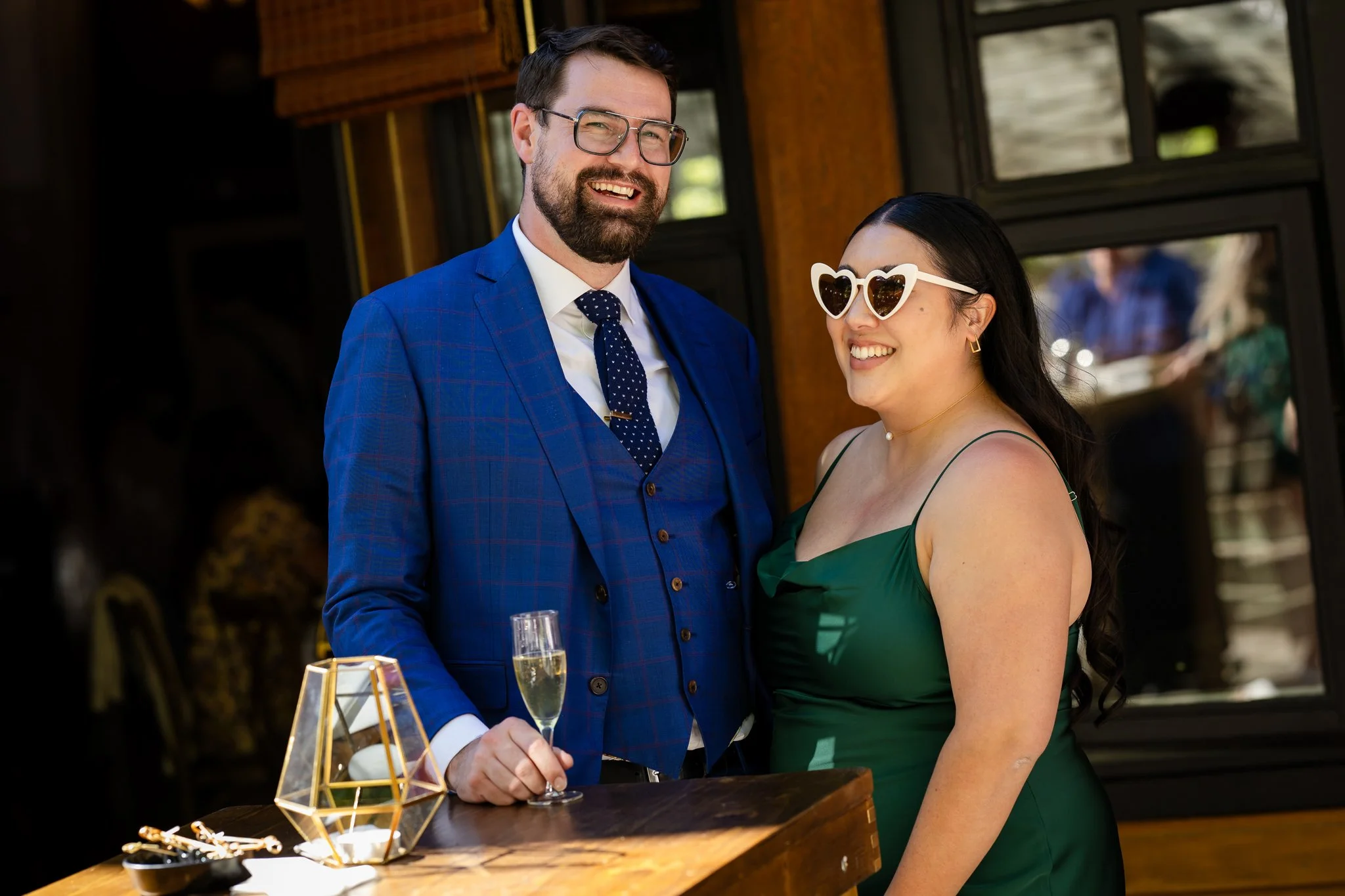 A man and woman at a social event, dressed in formal attire, smiling and holding glasses of champagne. The man is wearing a blue checkered suit and glasses, and the woman is wearing a green dress with heart-shaped sunglasses.