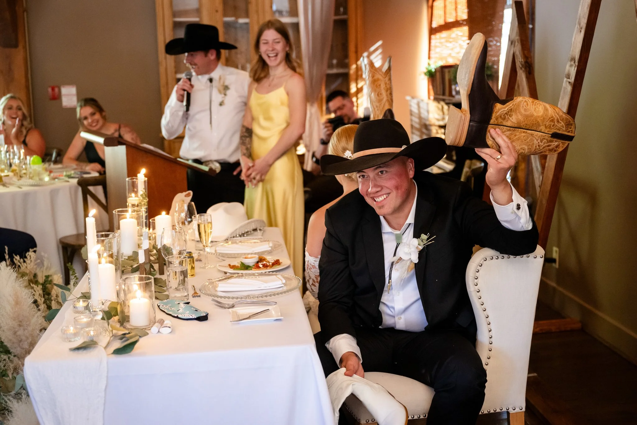 Man wearing a black cowboy hat and suit smiling and holding a cowboy boot over his head during a wedding reception, with women and a man in the background.
