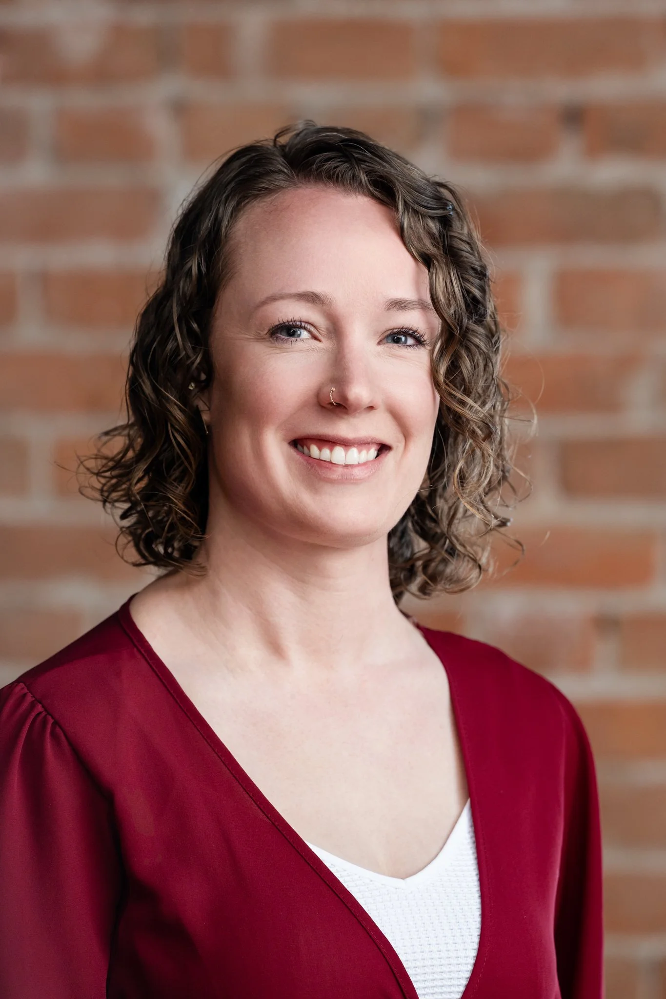 Portrait of a woman with curly brown hair, wearing a maroon top and a white inner shirt, smiling in front of a brick wall background.