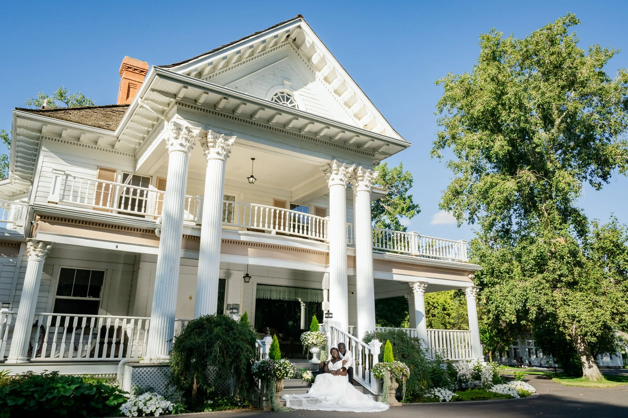 A white, multi-story house with large pillars and balconies, surrounded by greenery. Two people, dressed in wedding attire, sit on the front steps.