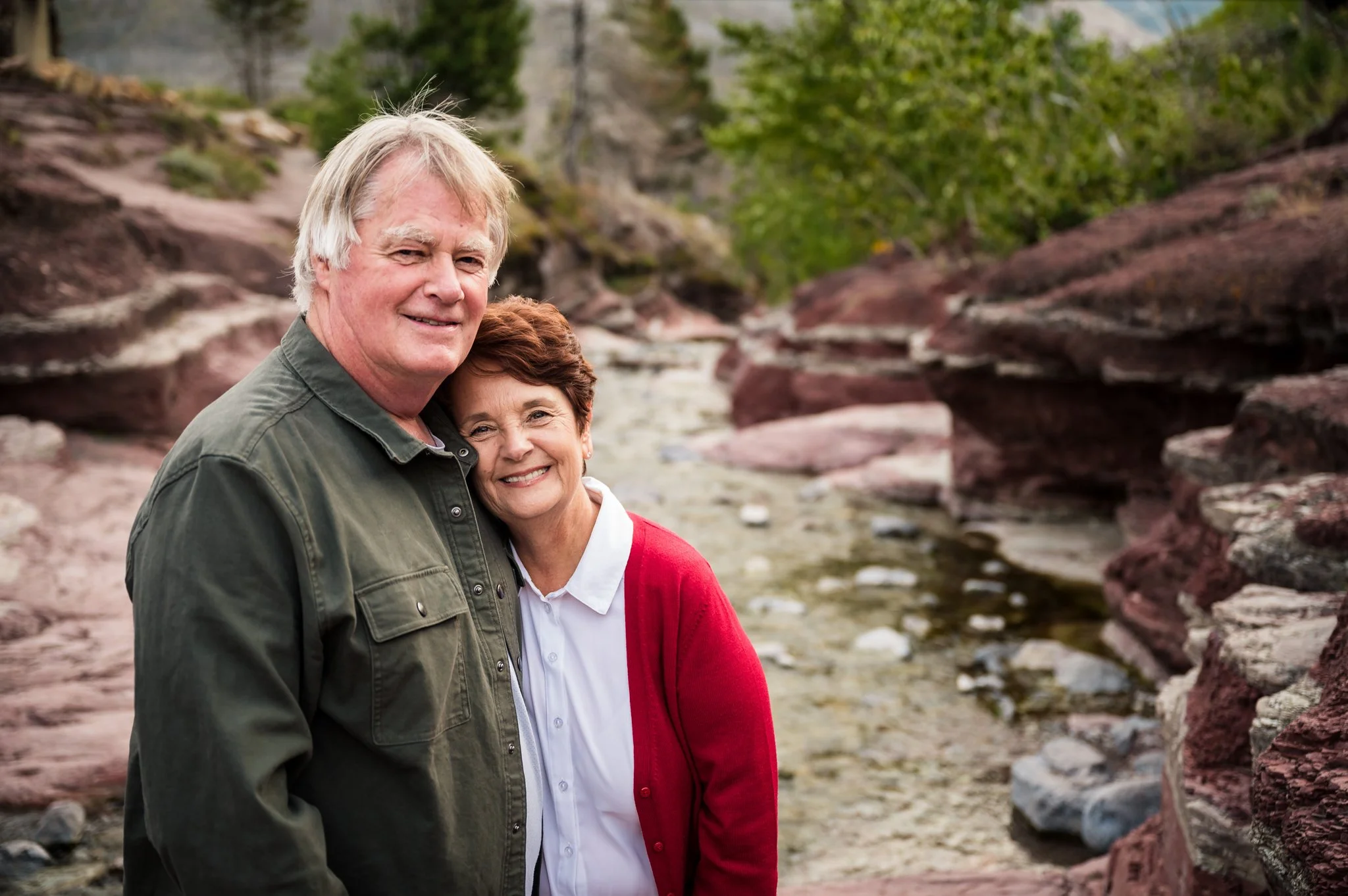 An elderly couple smiling and posing outdoors near a creek with rocks and green trees in the background.