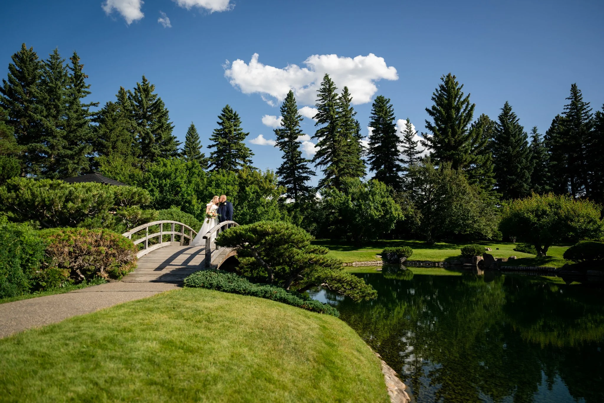 A bride and groom standing on a small arched bridge over a pond in a lush, green park, with tall trees and a blue sky with white clouds in the background.