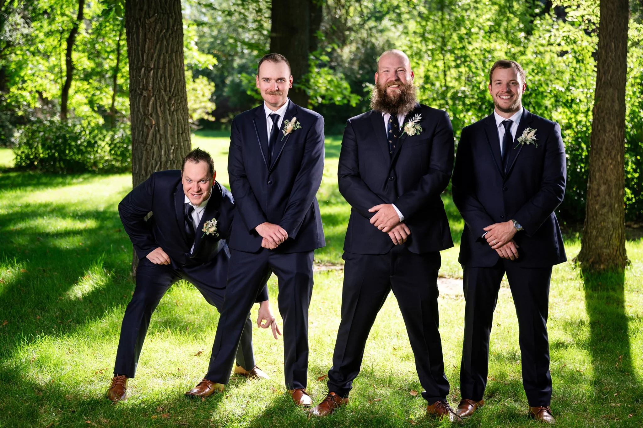 Five men dressed in dark suits standing outdoors in a grassy, wooded area for a wedding photo.
