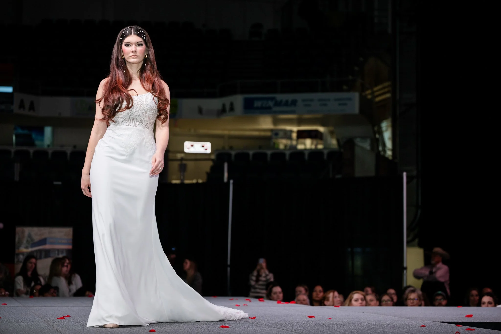A woman with long, wavy red hair and pearl hair accessories walking on a runway during a fashion show, wearing a white gown and surrounded by seated audience members.