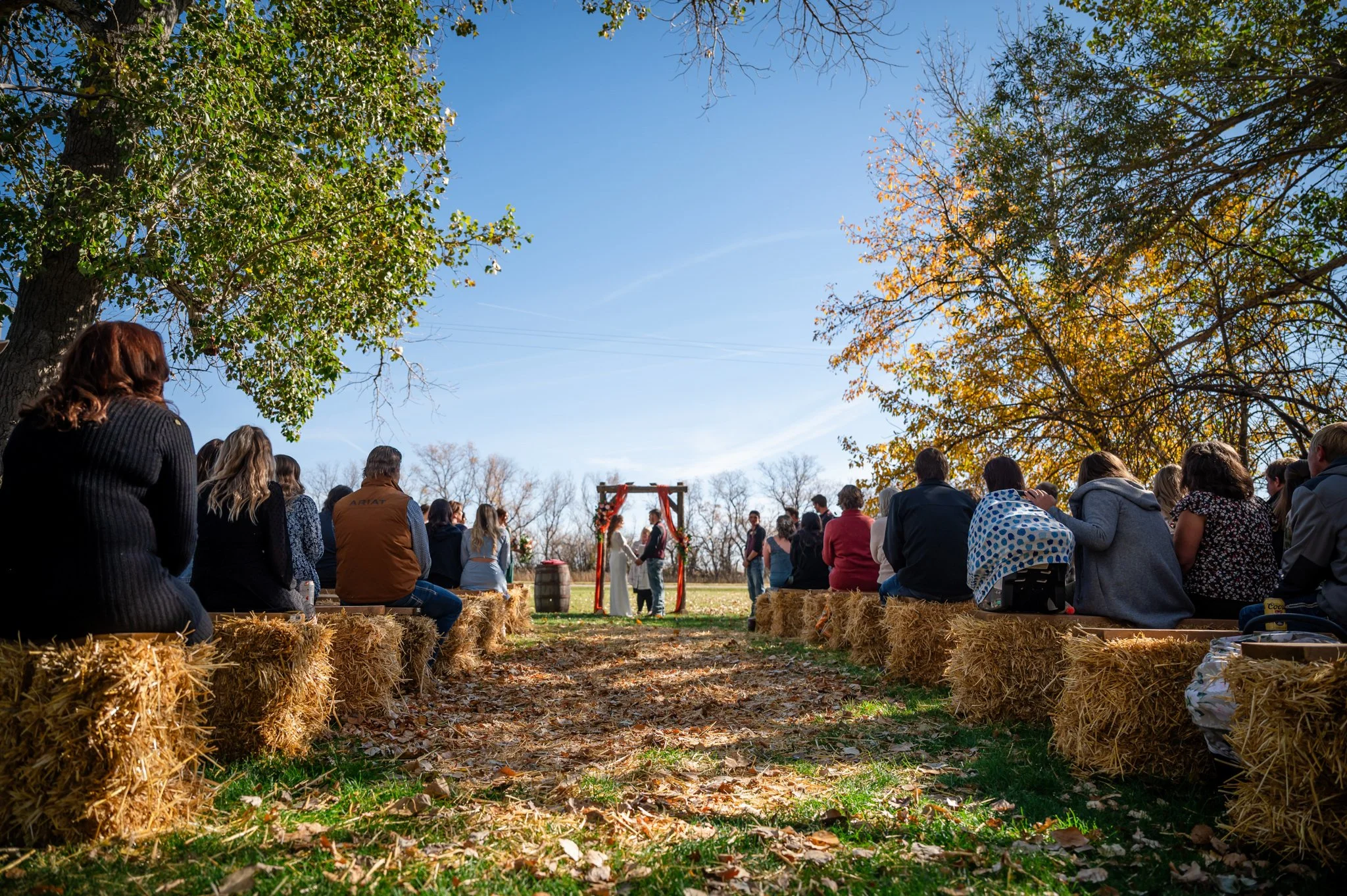 Outdoor wedding ceremony with guests seated on hay bales under trees on a sunny fall day.