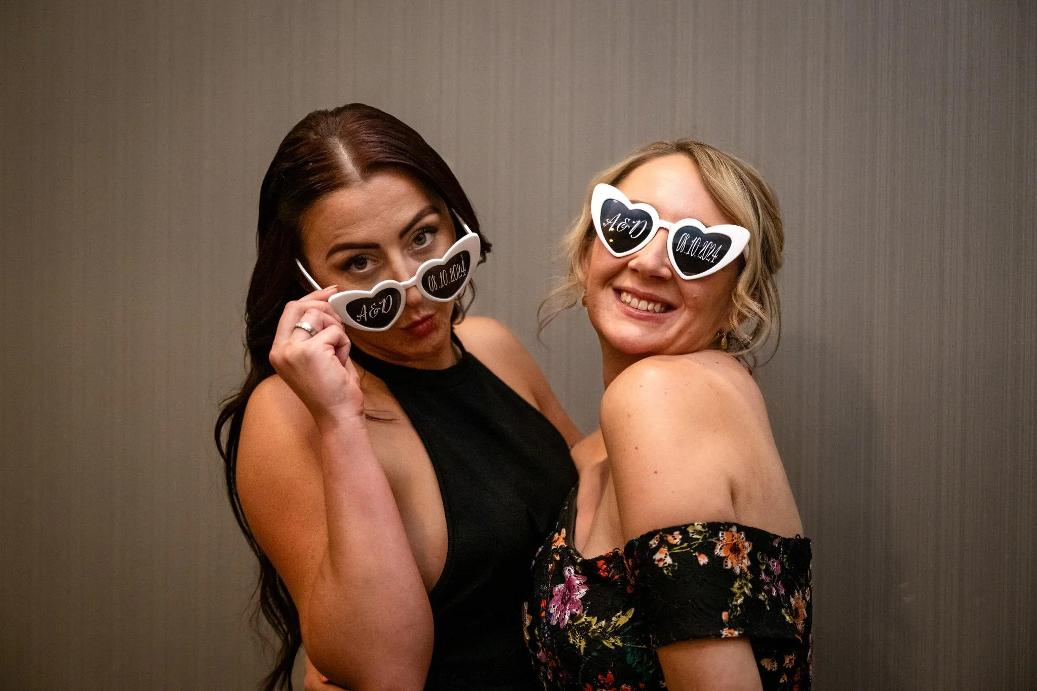 Two women wearing heart-shaped sunglasses smiling and posing together at an indoor event.