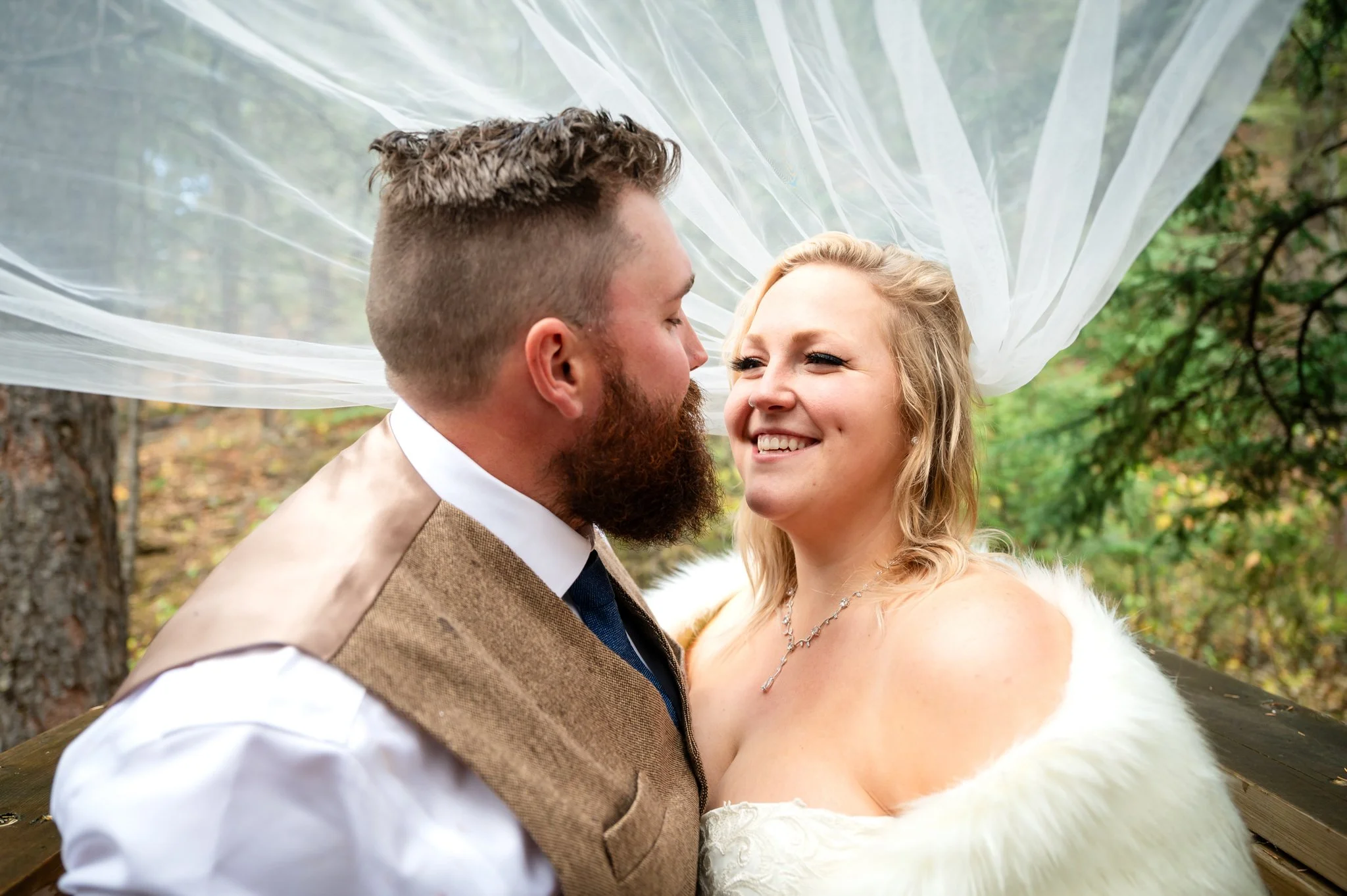 A bride and groom face each other smiling outdoors, with a white sheer fabric draped overhead and trees in the background.