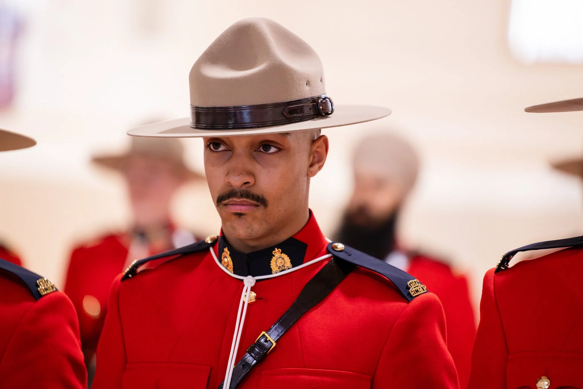 A Royal Canadian Mounted Police officer wearing a red uniform with black shoulder epaulets, a beige wide-brimmed hat with a black band, and a black strap across his chest. He has a serious expression and a mustache. Several other officers in similar
