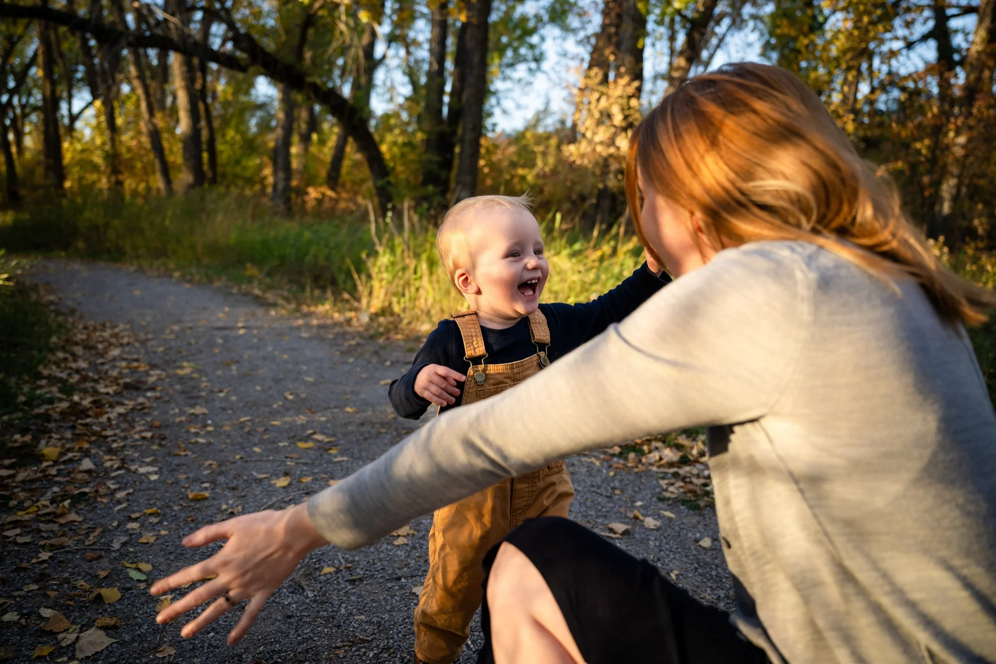 A woman and a smiling young boy playing on a wooded trail during autumn, with yellow fallen leaves on the ground and trees with autumn foliage in the background.