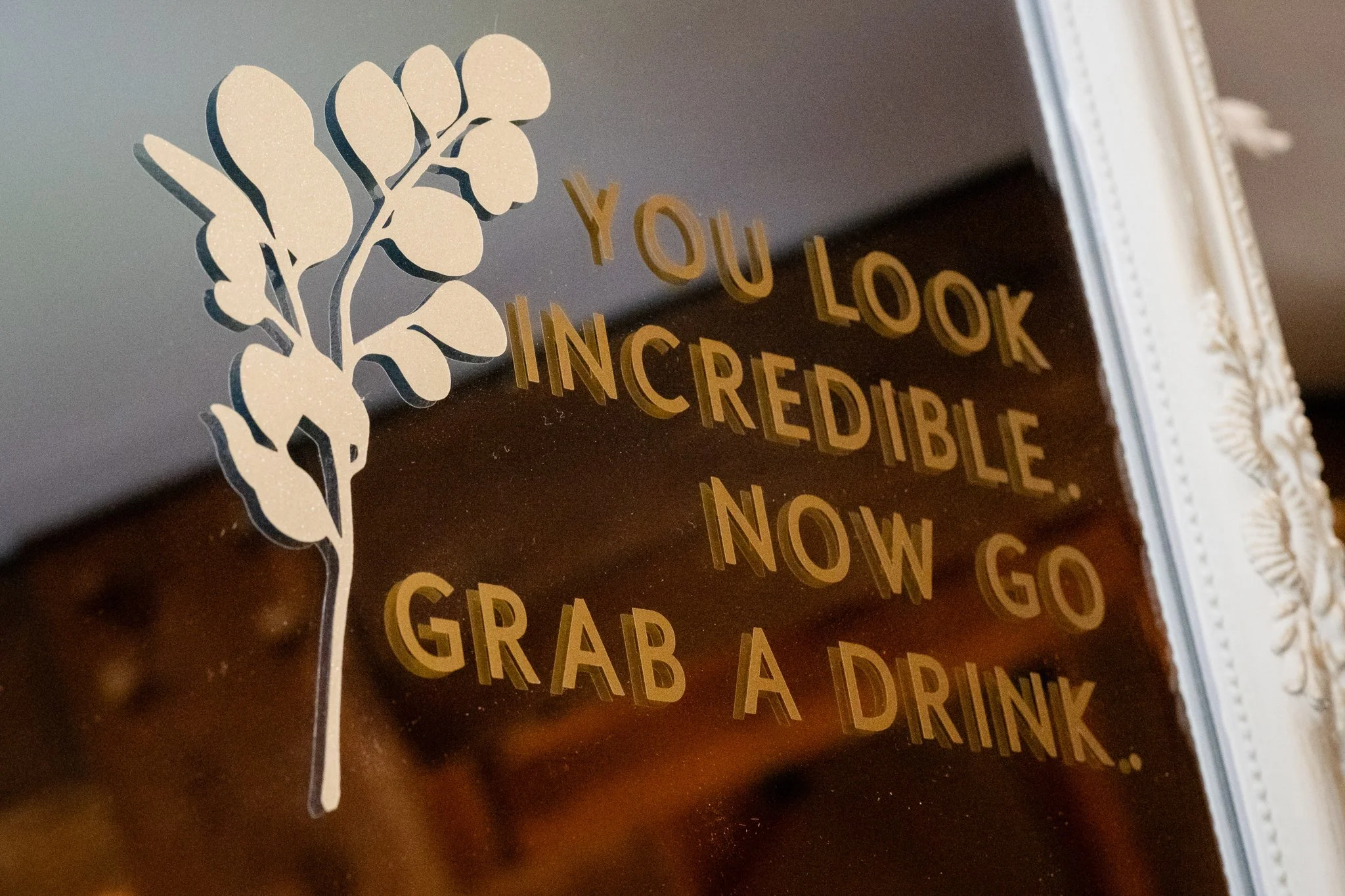 A decorative mirror with gold lettering that reads, "You look incredible. Now go grab a drink," and a white floral design on the top right edge.