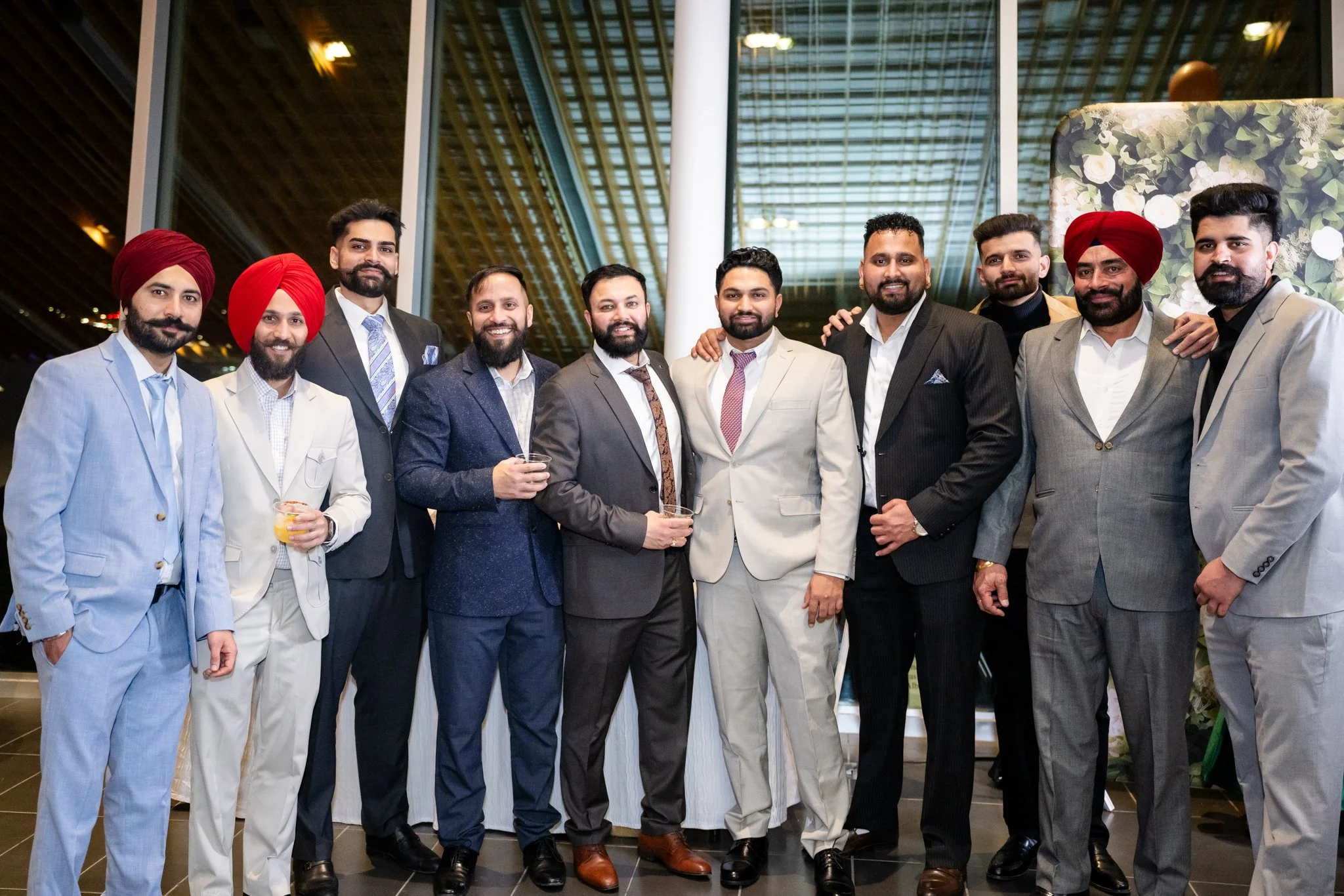 Group of men in suits at a formal event, some wearing turbans and holding drinks, standing in front of a large modern window with a decorative backdrop.