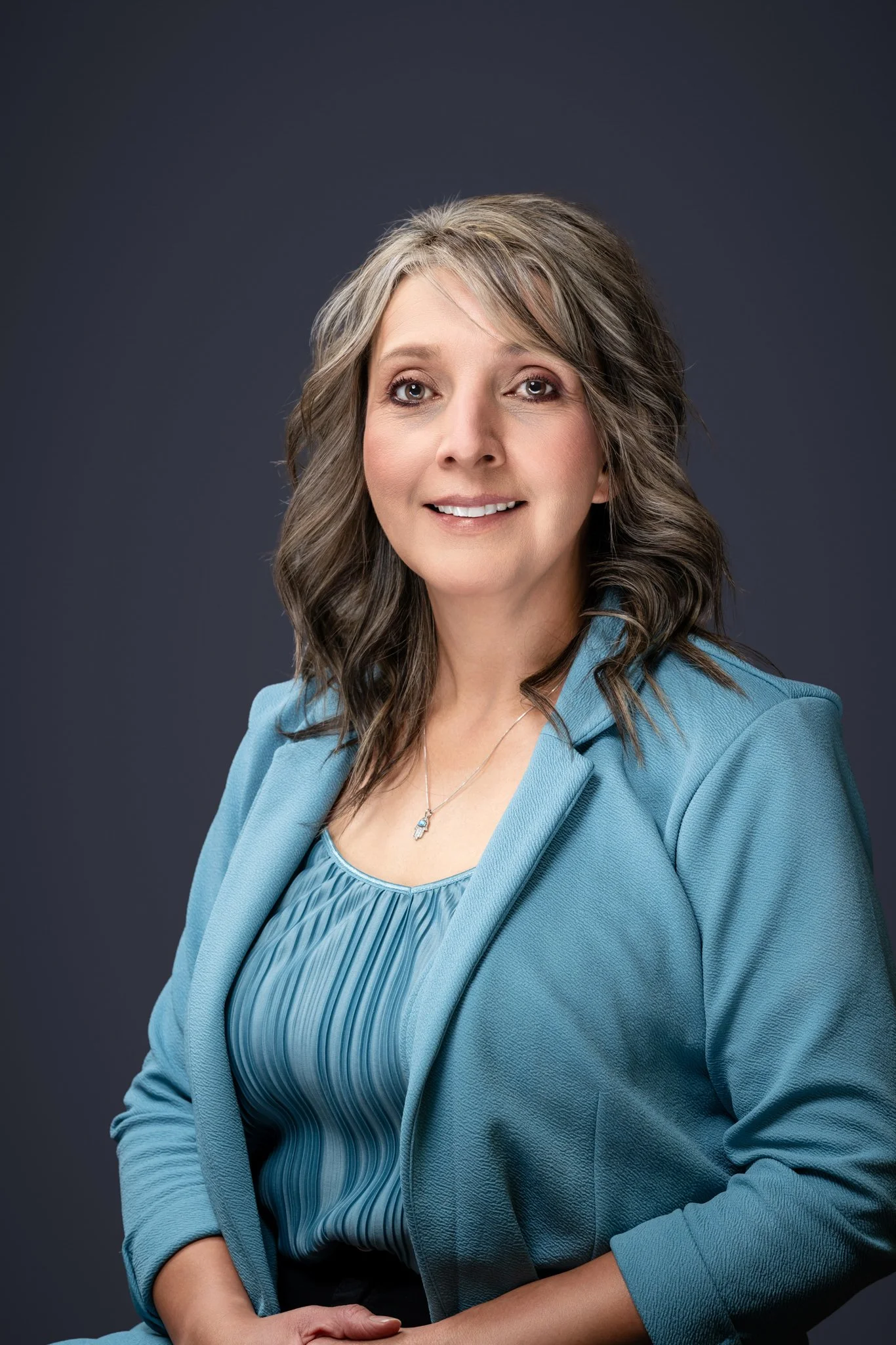 A woman with shoulder-length grayish-brown hair in loose waves, wearing a blue blazer over a matching blouse, smiling softly against a dark gray background.