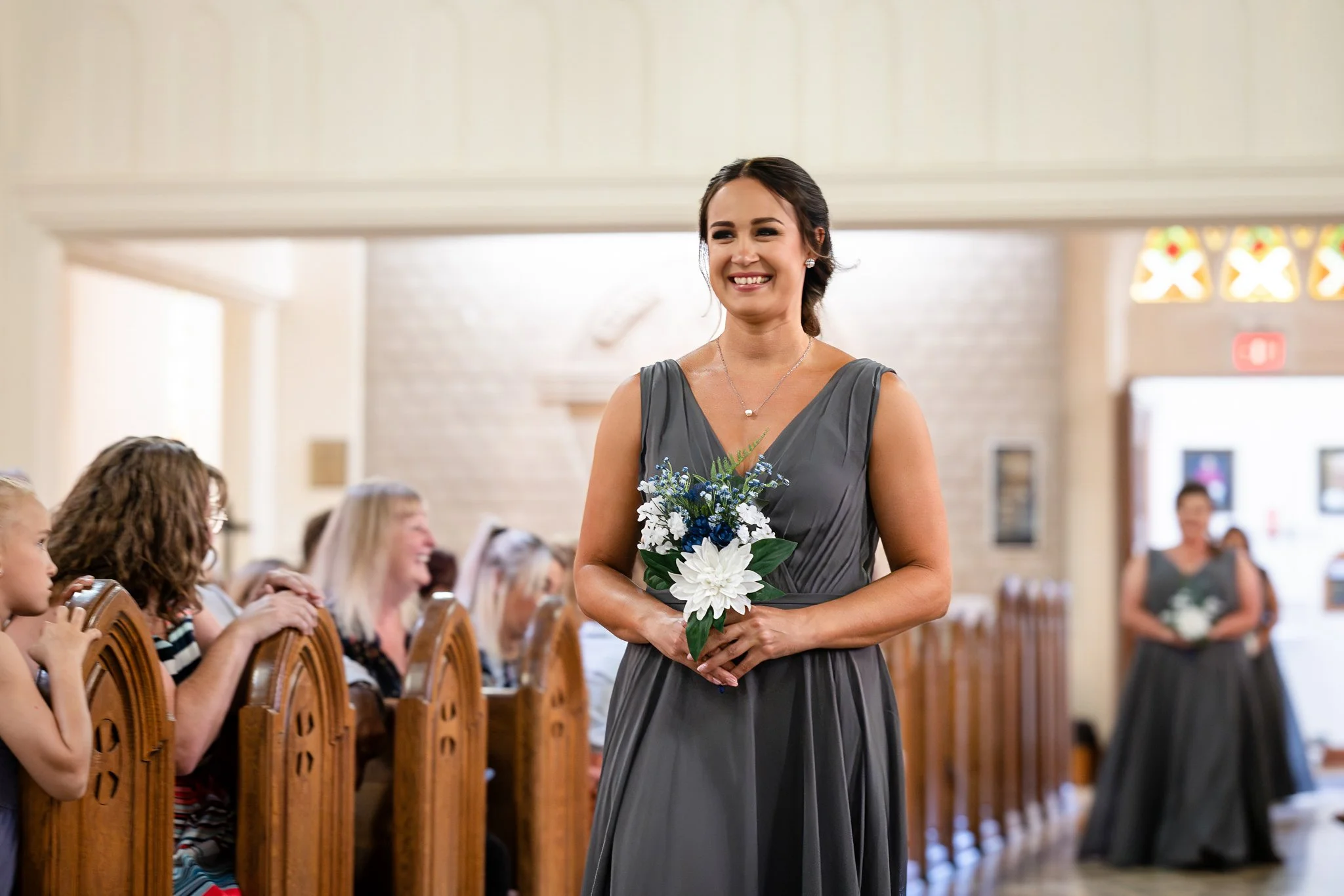 A smiling woman in a gray dress holding a bouquet at a wedding ceremony inside a church.