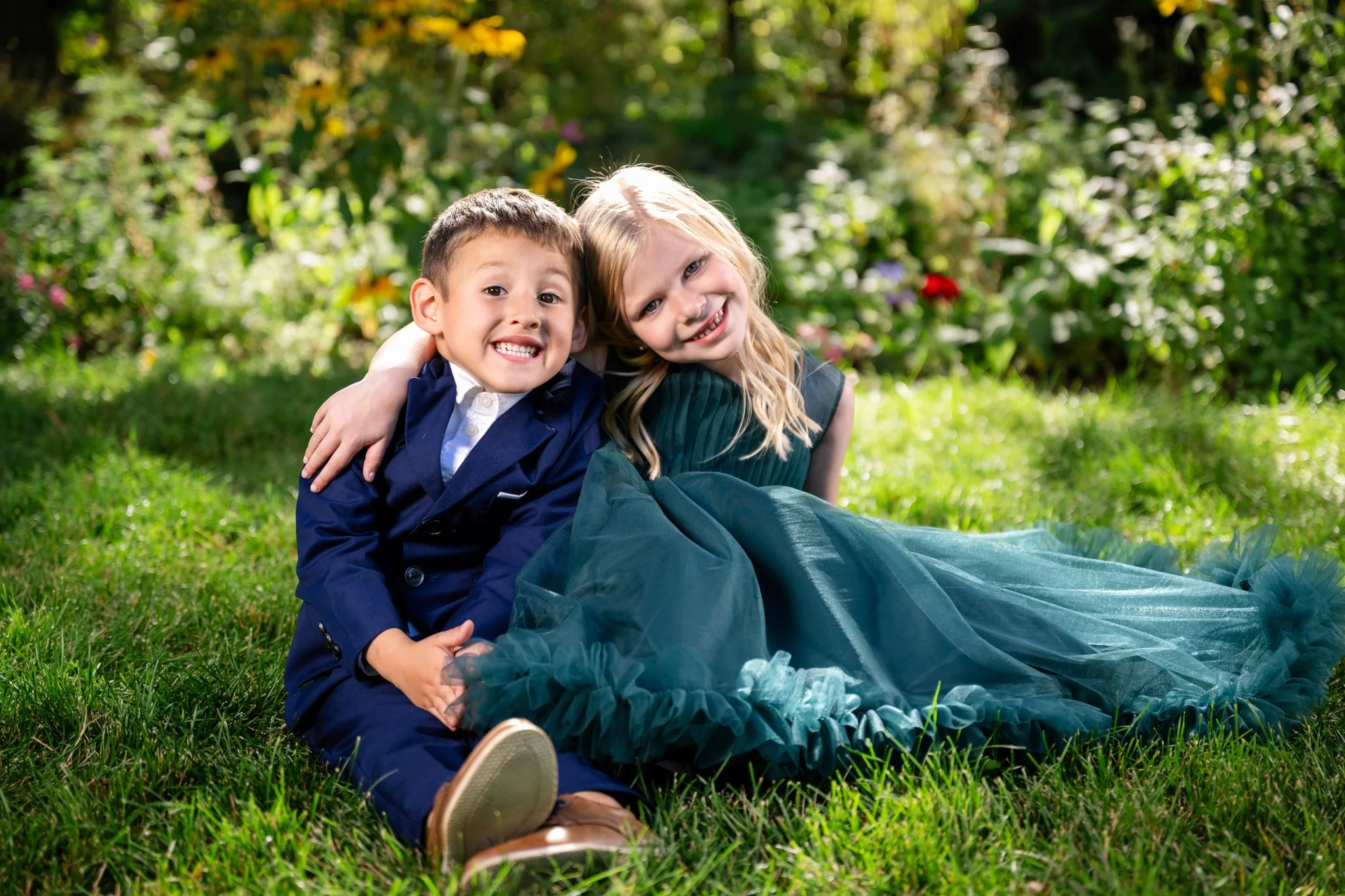 Two children, a boy and a girl, sitting on grass in a garden, smiling and leaning on each other.