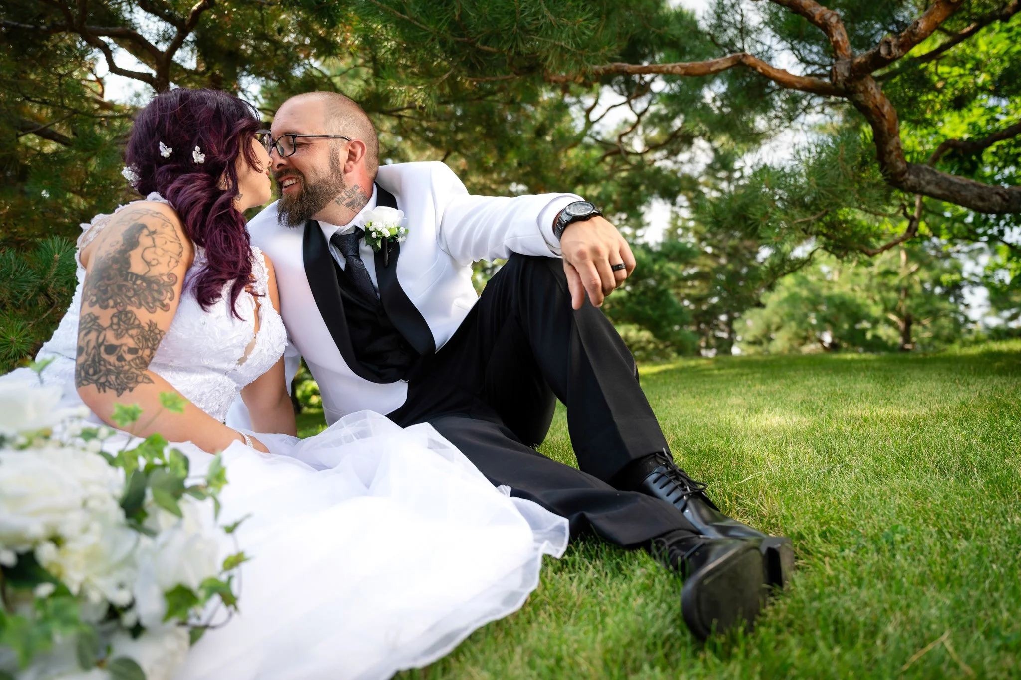 A newlywed couple sitting on grass under a tree, leaning close together, smiling and touching noses. The bride has purple hair with floral accessories, tattoos on her arm, and is wearing a white wedding dress. The groom has a beard, glasses, and is d