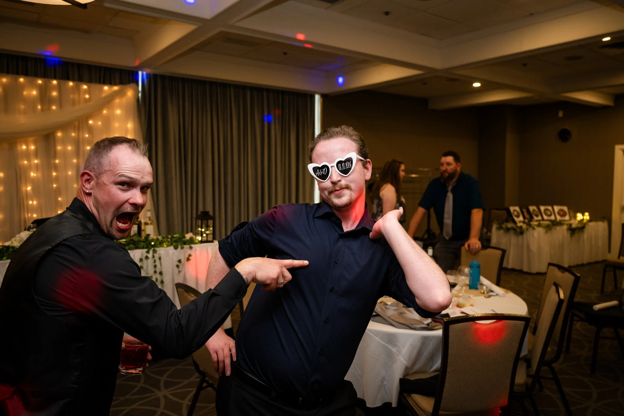 Two men celebrating at a party, one pointing at the other who is wearing sunglasses that say "40" and the date "09.10.22" in a decorated event hall.