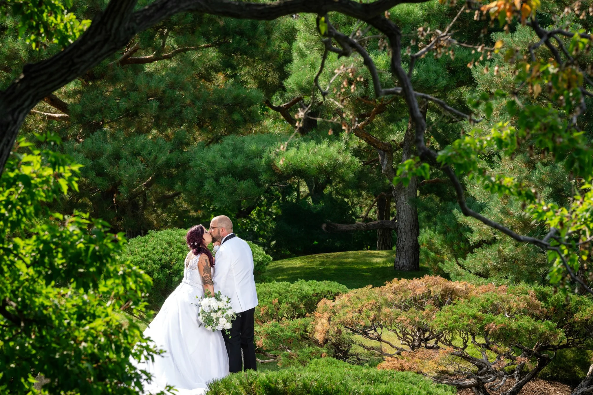 A bride and groom share a kiss in a lush, green garden surrounded by trees and shrubs.