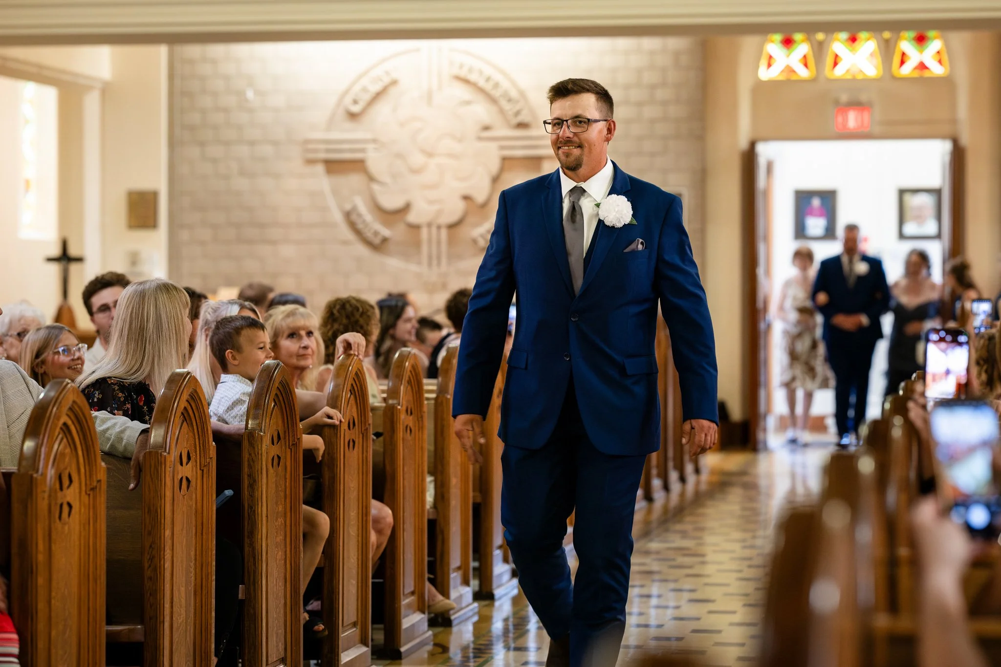 Man in a blue suit walking down the aisle at a wedding, with guests seated on either side.