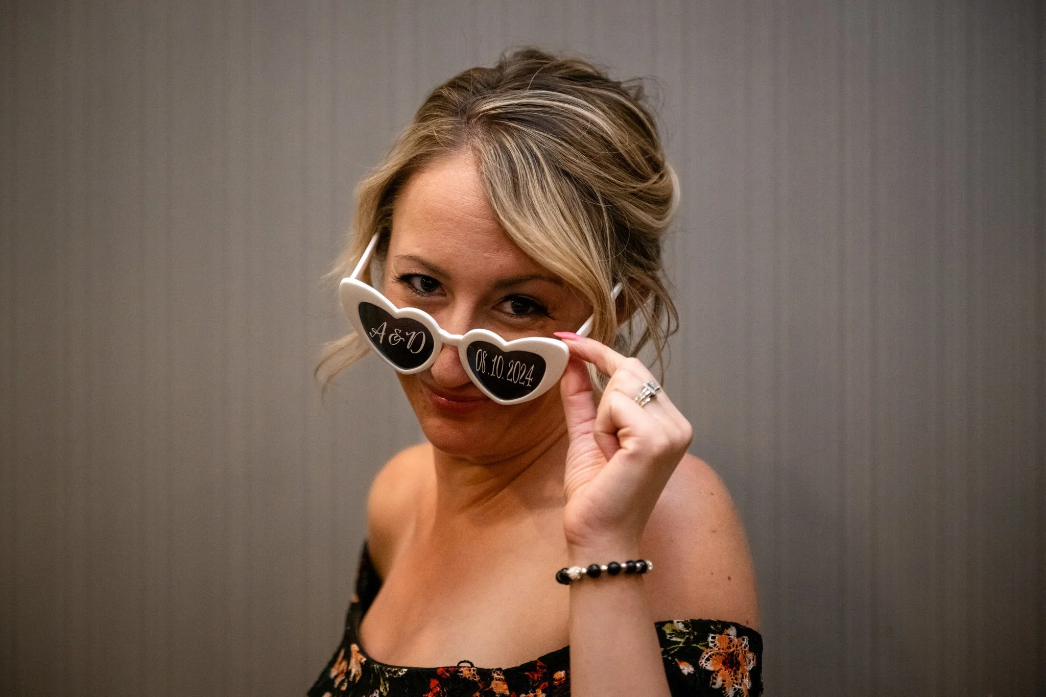A woman with blonde hair wearing sunglasses with the initials 'A&D' and a date '08.10.2024', smiling and looking at the camera, holding the sunglasses slightly away from her face.