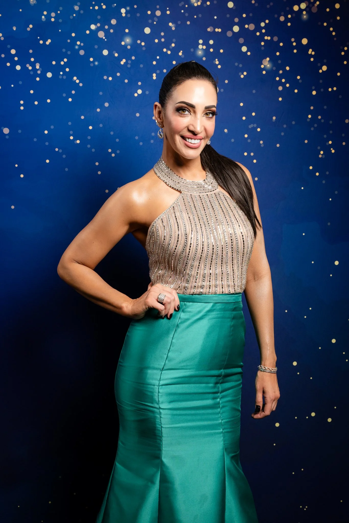 A woman in a sparkling beige top and green skirt standing against a dark blue background with gold confetti-like patterns, smiling and posing with one hand on her hip.