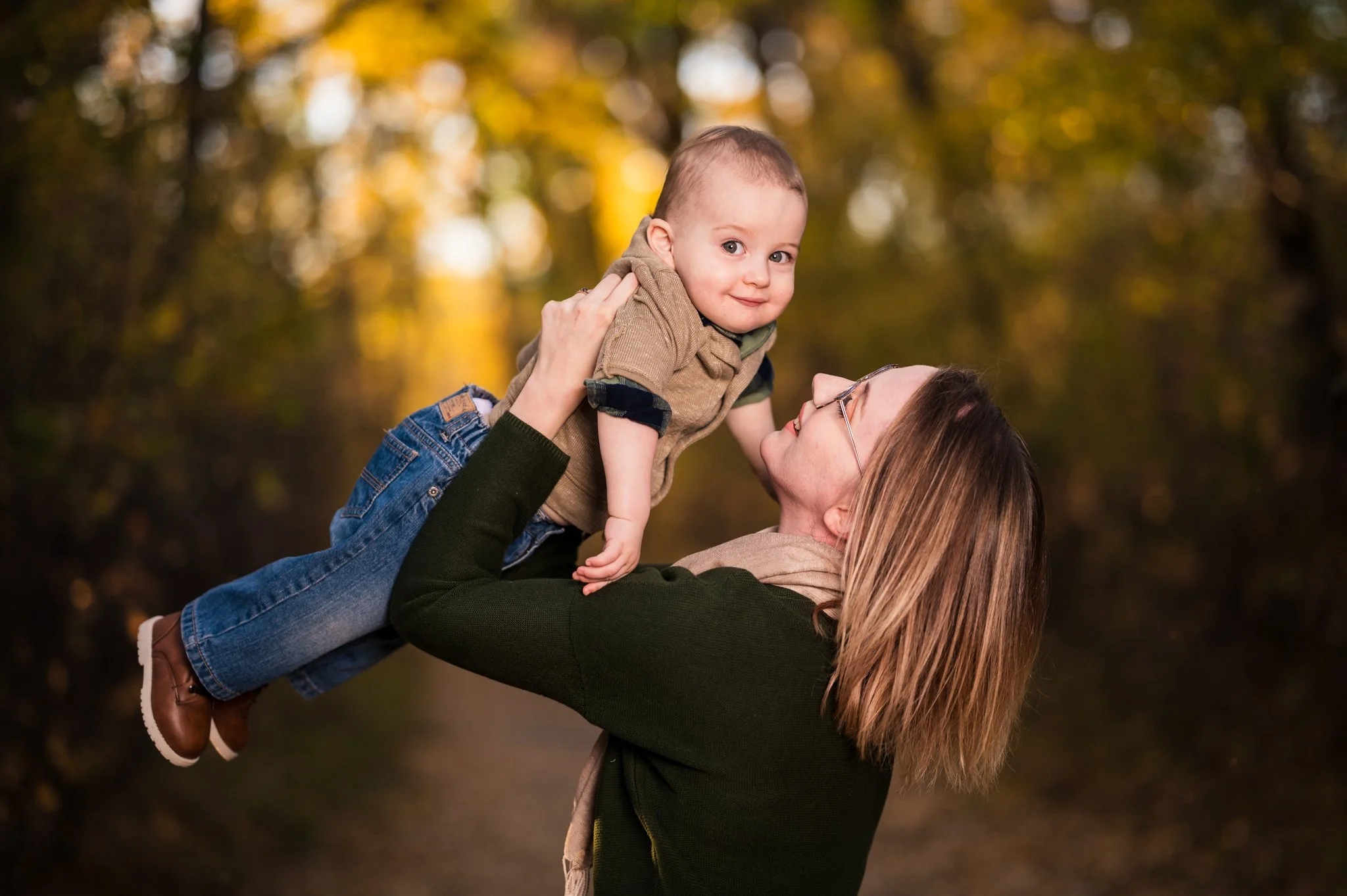A woman with shoulder-length brown hair and glasses holding a smiling young boy with short hair in a wooded autumn setting.