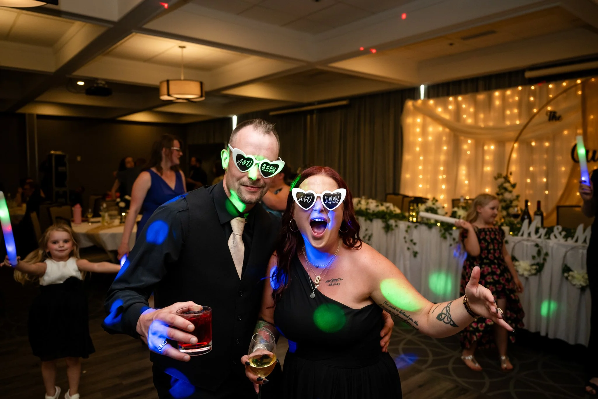 Two people celebrating at a wedding reception, both wearing novelty sunglasses with hearts and initials, holding drinks, smiling and enjoying the party.