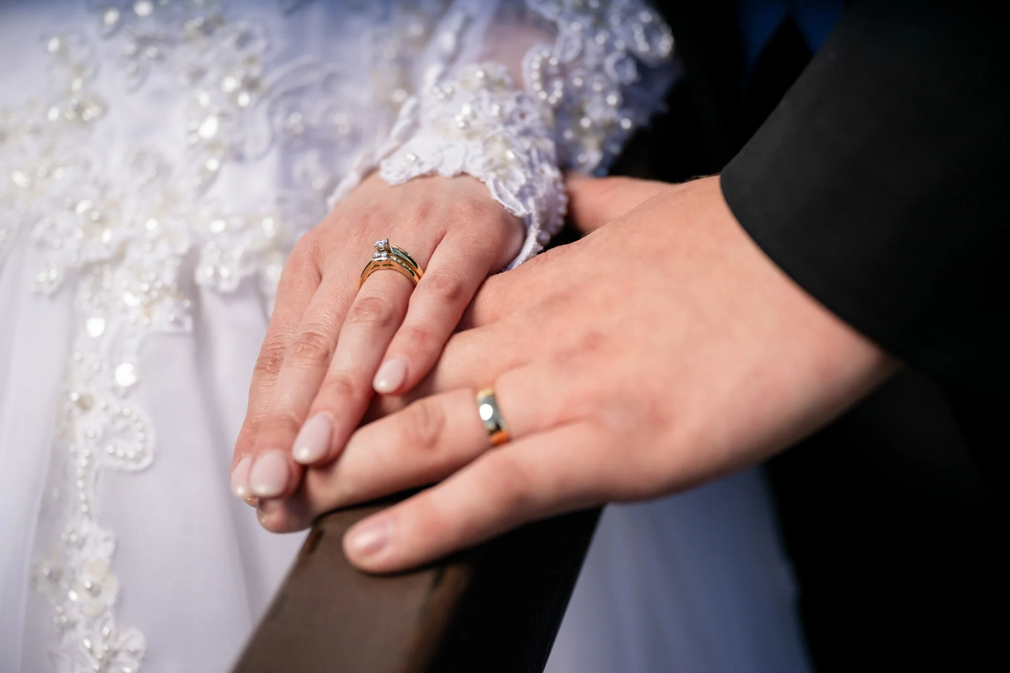 Close-up of a bride and groom holding hands, showing wedding rings. The bride's hand has a wedding ring with a diamond, and the groom's hand has a simple band. The bride's attire is embroidered with lace and beads.