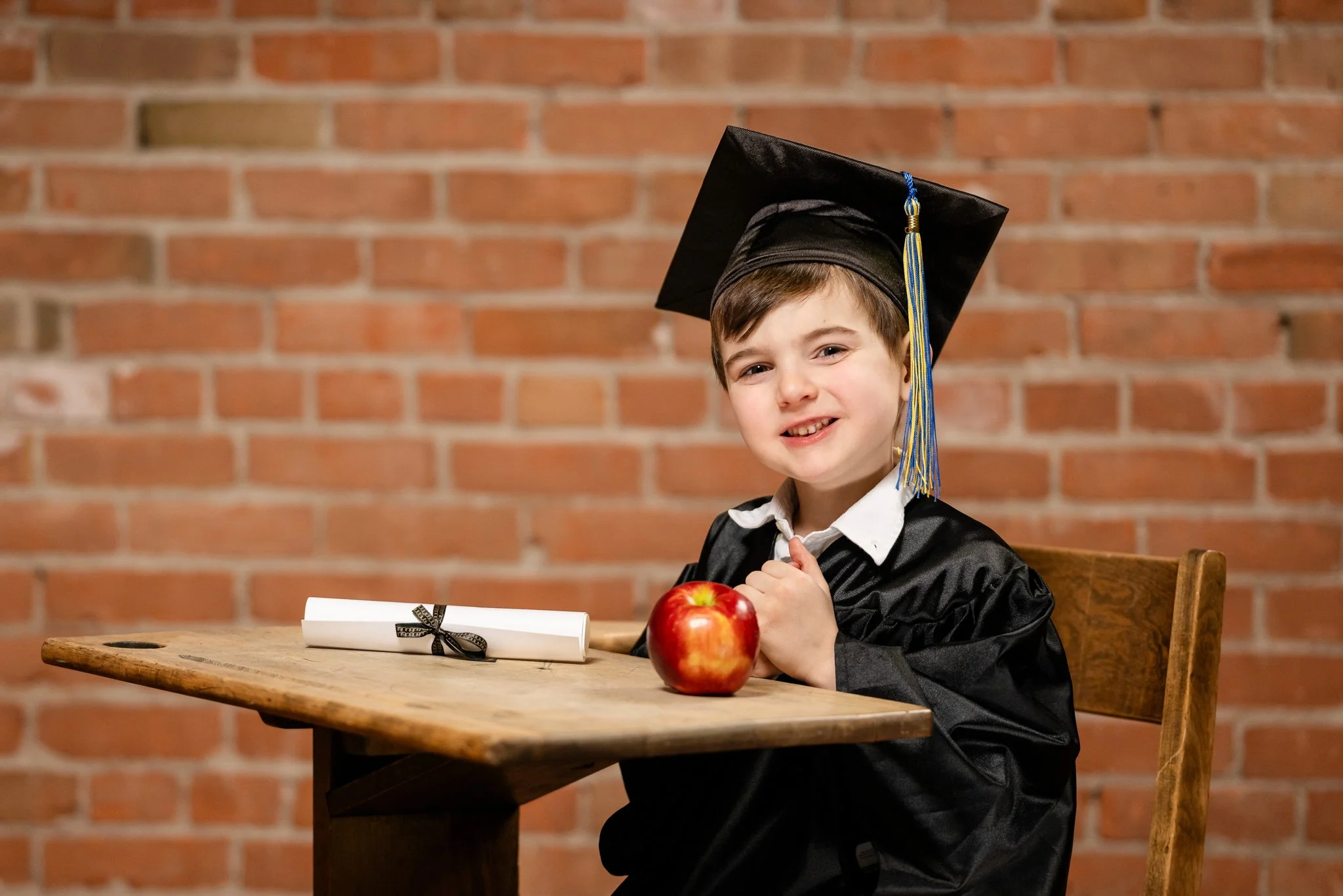 A young boy in a graduation cap and gown sitting at a wooden table with a rolled diploma tied with a ribbon and a red apple, smiling against a brick wall background.