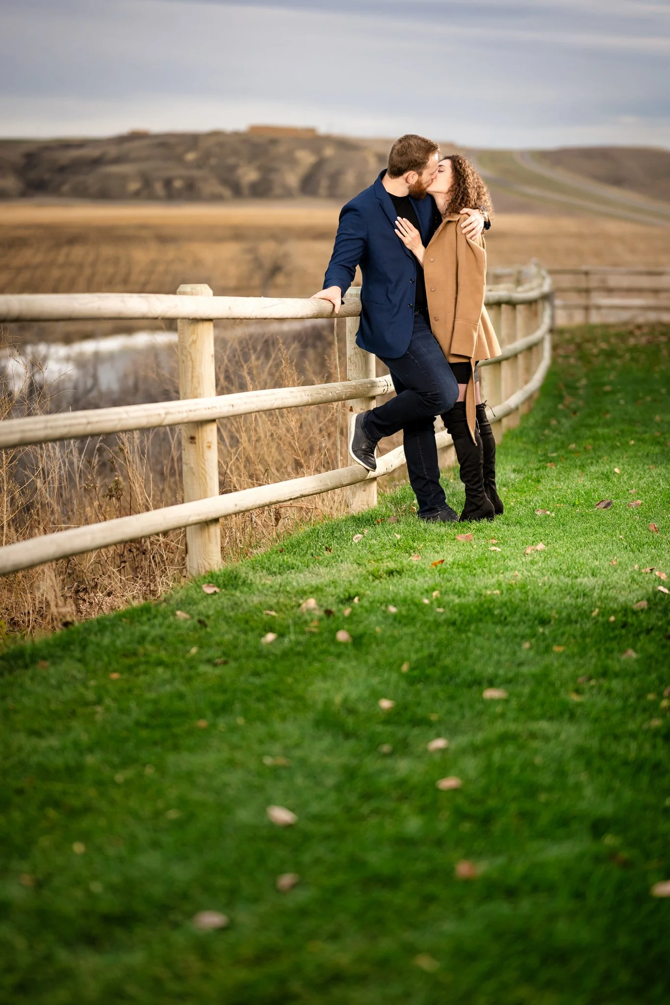 A couple sharing a kiss outdoors on a grassy area near a wooden fence, with a hilly landscape in the background.