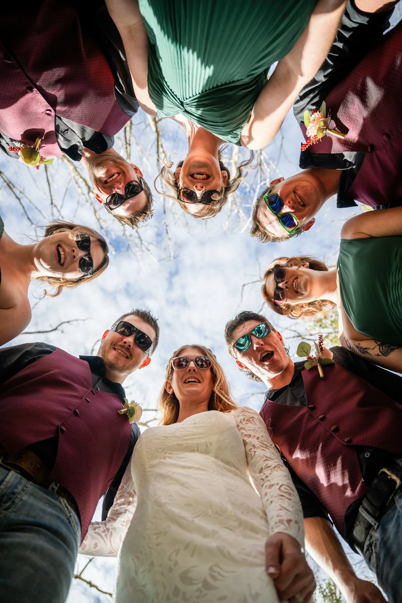 Group of people in a circle looking down at the camera, smiling, taken outdoors with a blue sky and tree branches in the background.