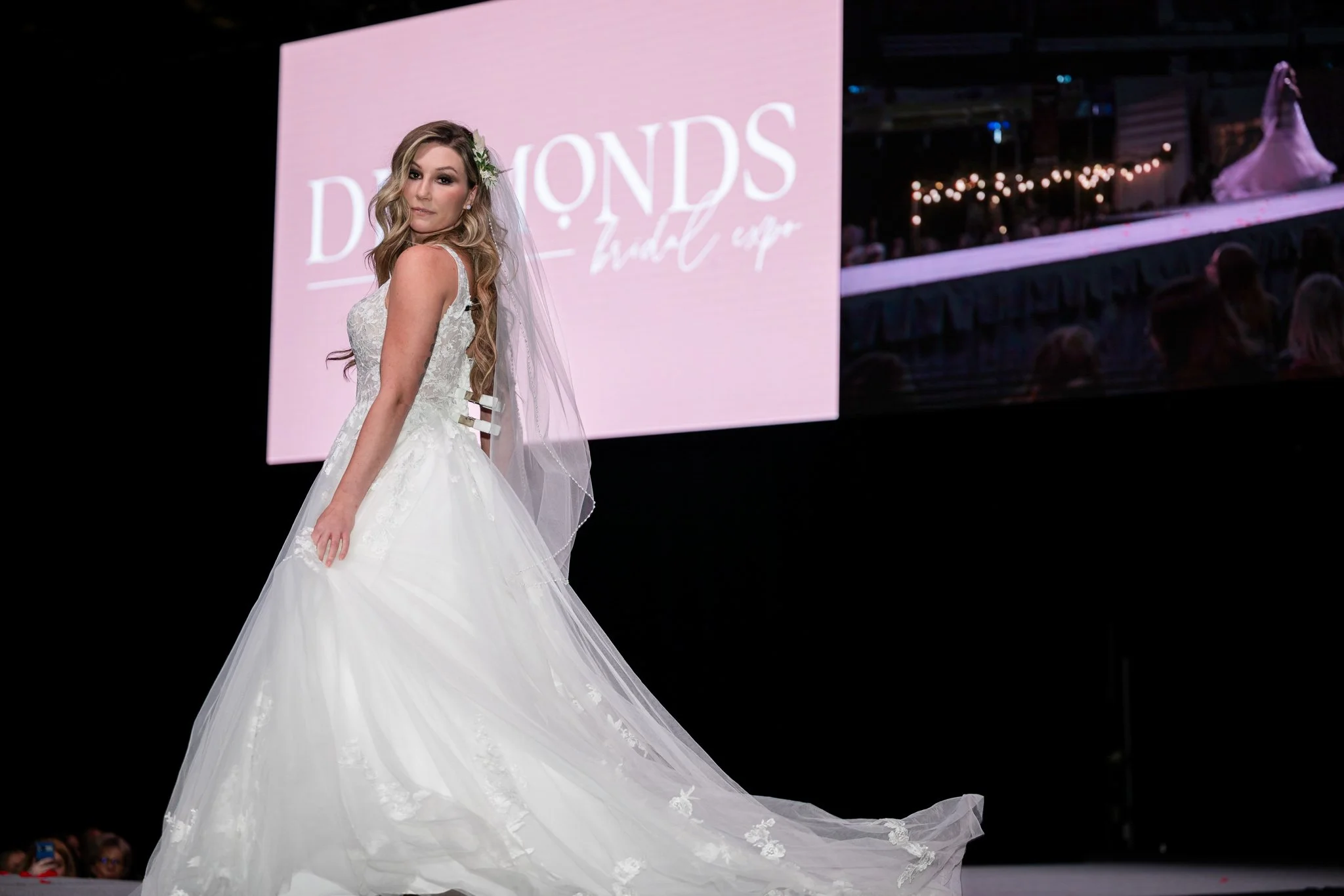 A woman in a white wedding dress on a runway, with a pink sign in the background reading 'Diamonds Bridal Expo'.