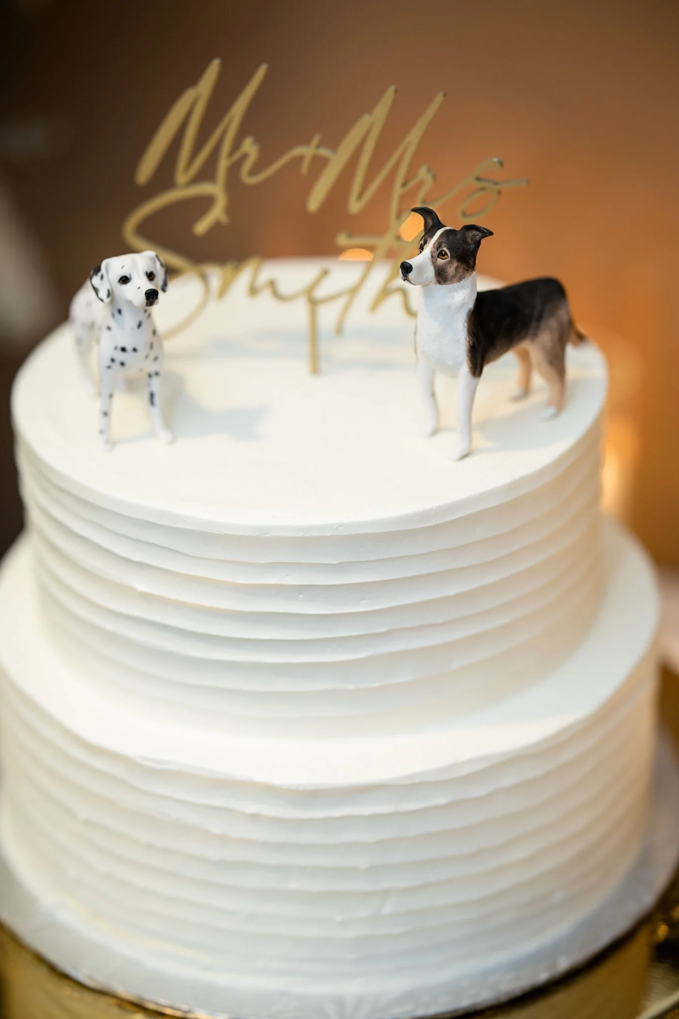 White frosted cake with two dog figurines, a Dalmatian and a Border Collie, as cake toppers, and a gold 'Mr & Mrs' cake topper in the background.