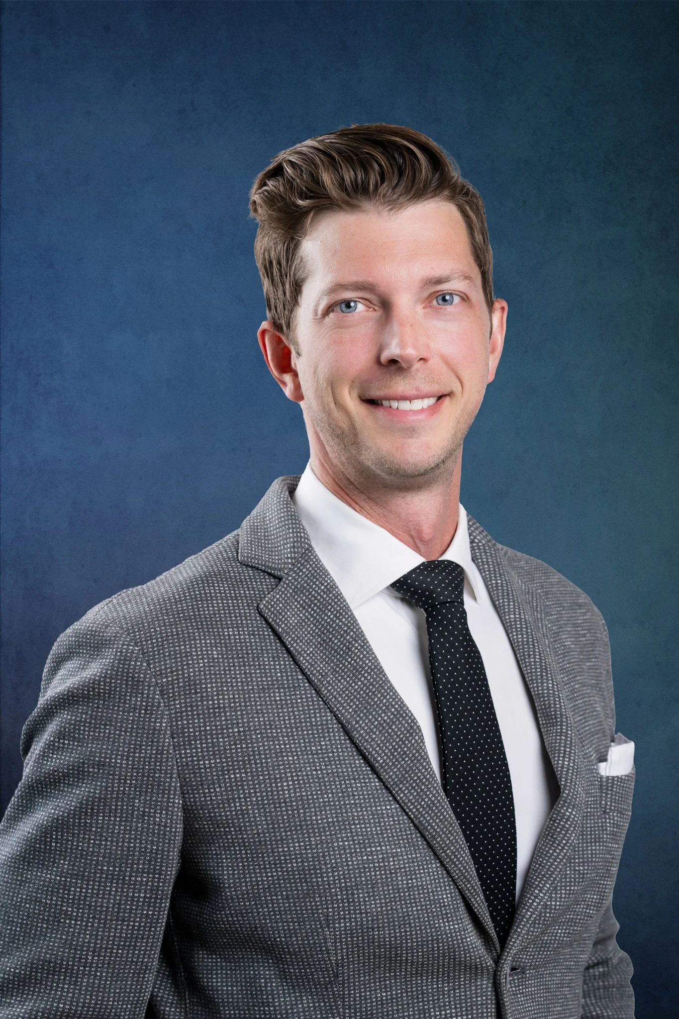 Portrait of a young man wearing a gray checked suit, white shirt, and black polka dot tie, smiling against a blue background.