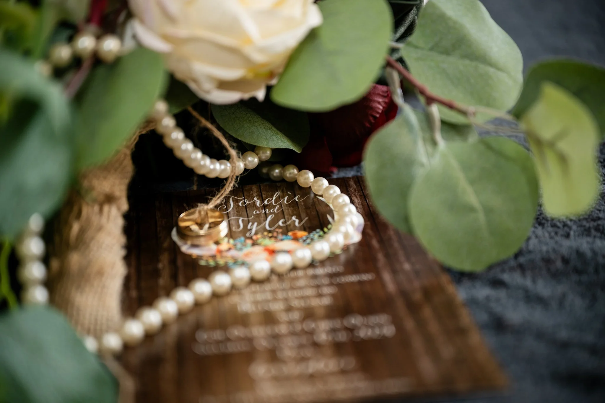 A close-up of wedding rings, pearls, greenery, and a sign with the names 'Tordie and Tyler' on a wooden surface.