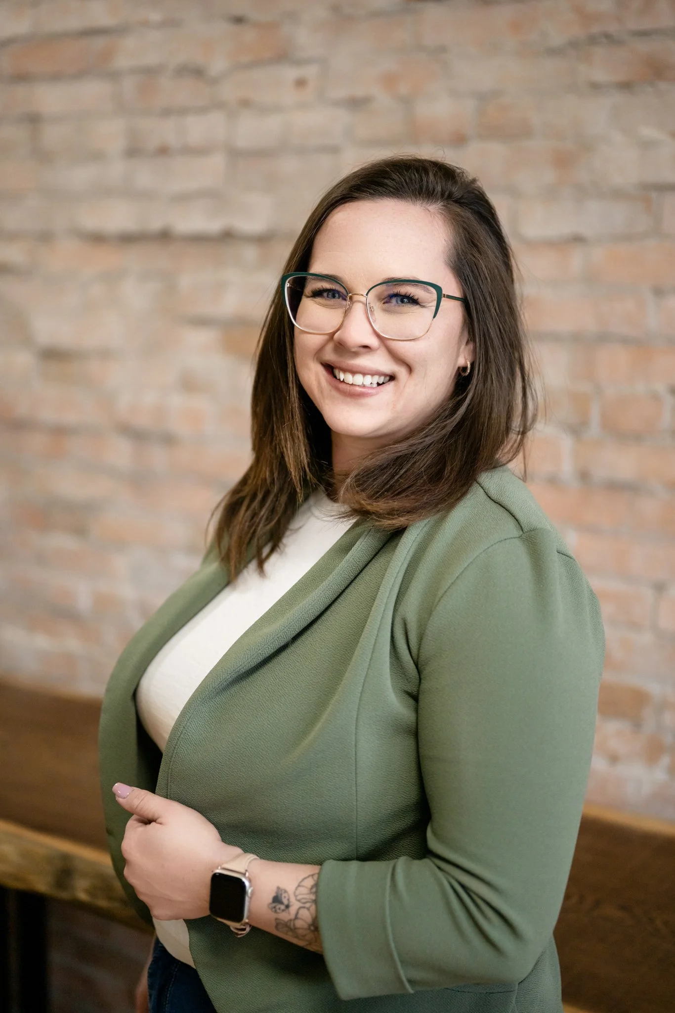A woman with brown hair, glasses, and a green blazer smiling indoors against a brick wall.