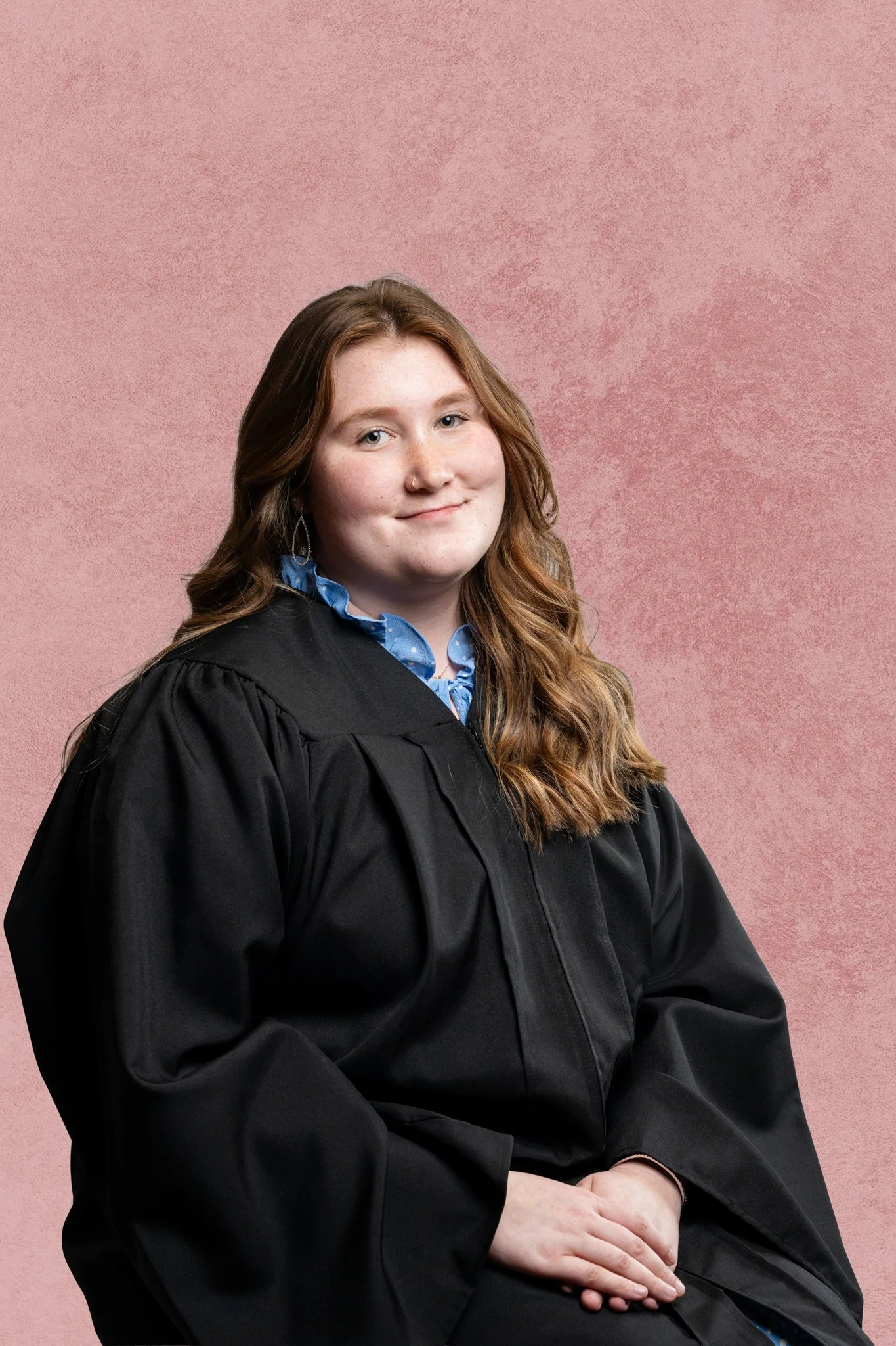 A young woman with long wavy reddish-brown hair, wearing a graduation cap and gown, sitting against a pink textured background.