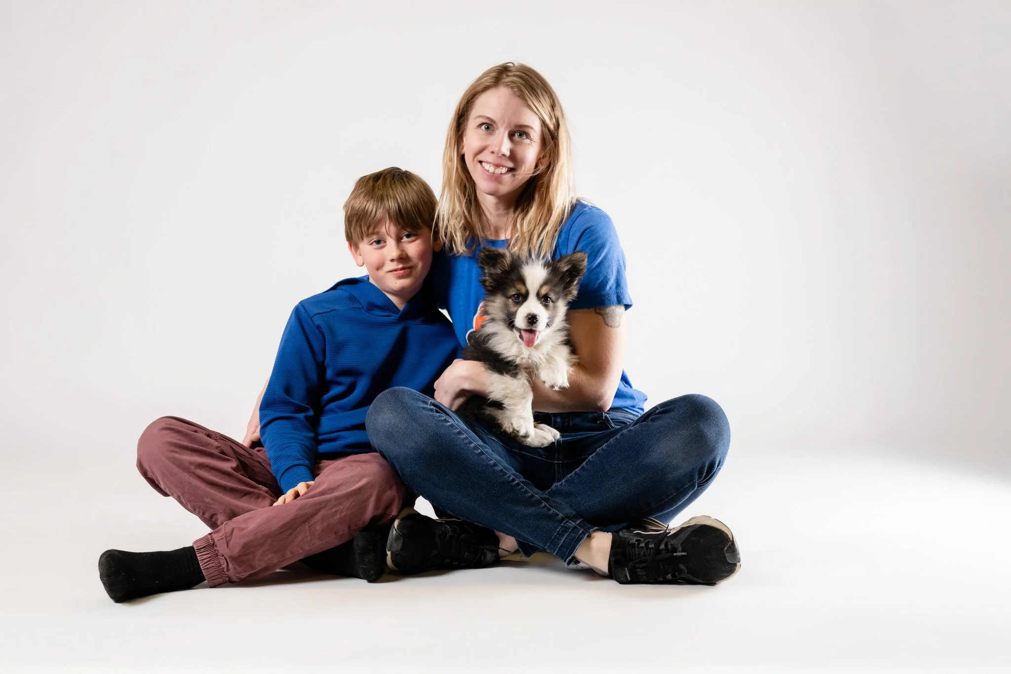 A woman and a young boy sitting on the floor with a puppy, all smiling, against a plain white background.