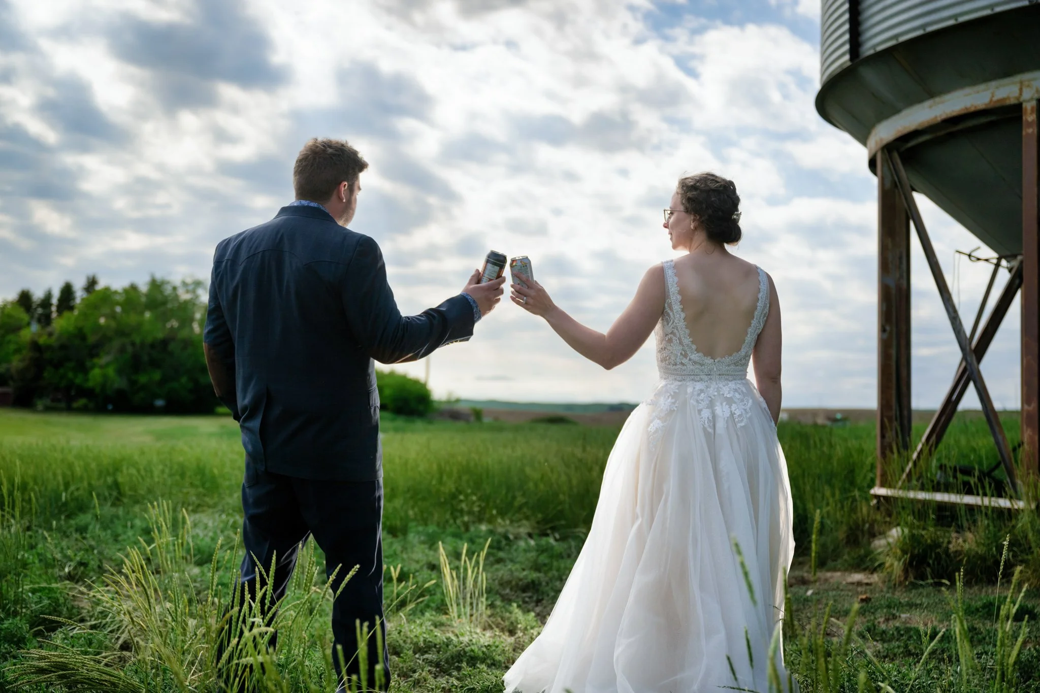 Bride and groom in a field raising drinks in a toast on a cloudy day, with green grass and trees in the background.