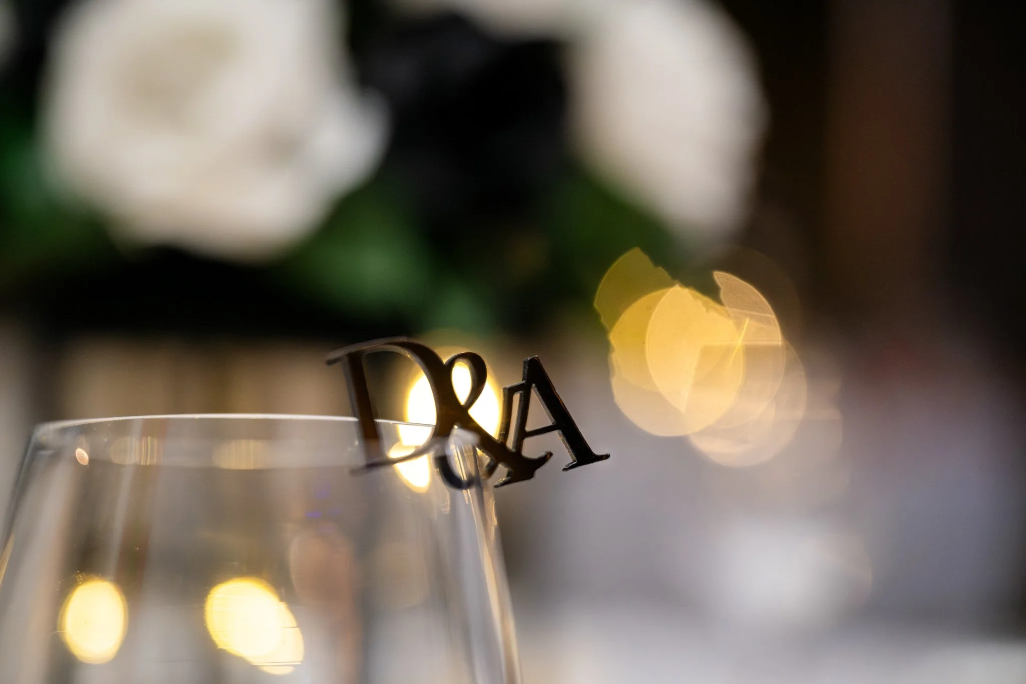 Close-up of a glass with a 'D&A' cake topper, blurred background with lights and flowers.