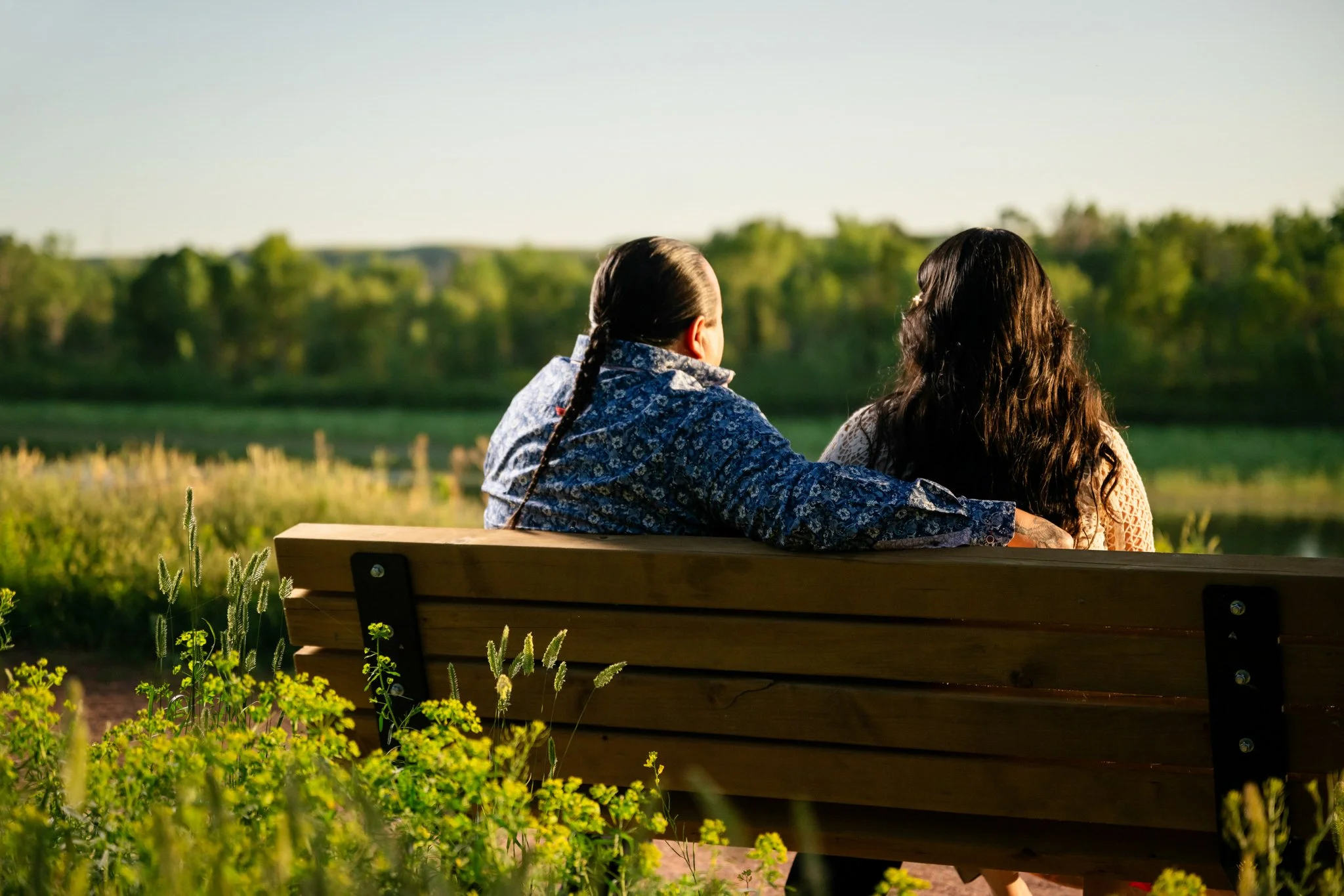 Two women sitting on a wooden bench outdoors, facing away, overlooking a river and lush green landscape in the background.