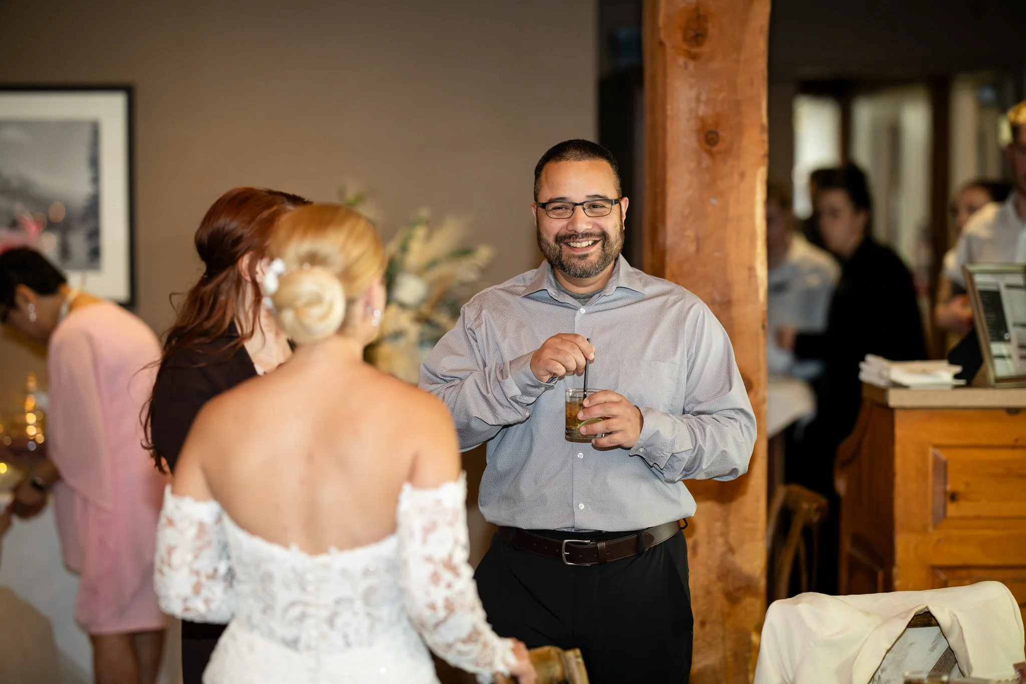 A man with glasses and a beard smiling while holding a drink with a straw, talking to two women, one wearing a white lace dress, at a social gathering or wedding reception.