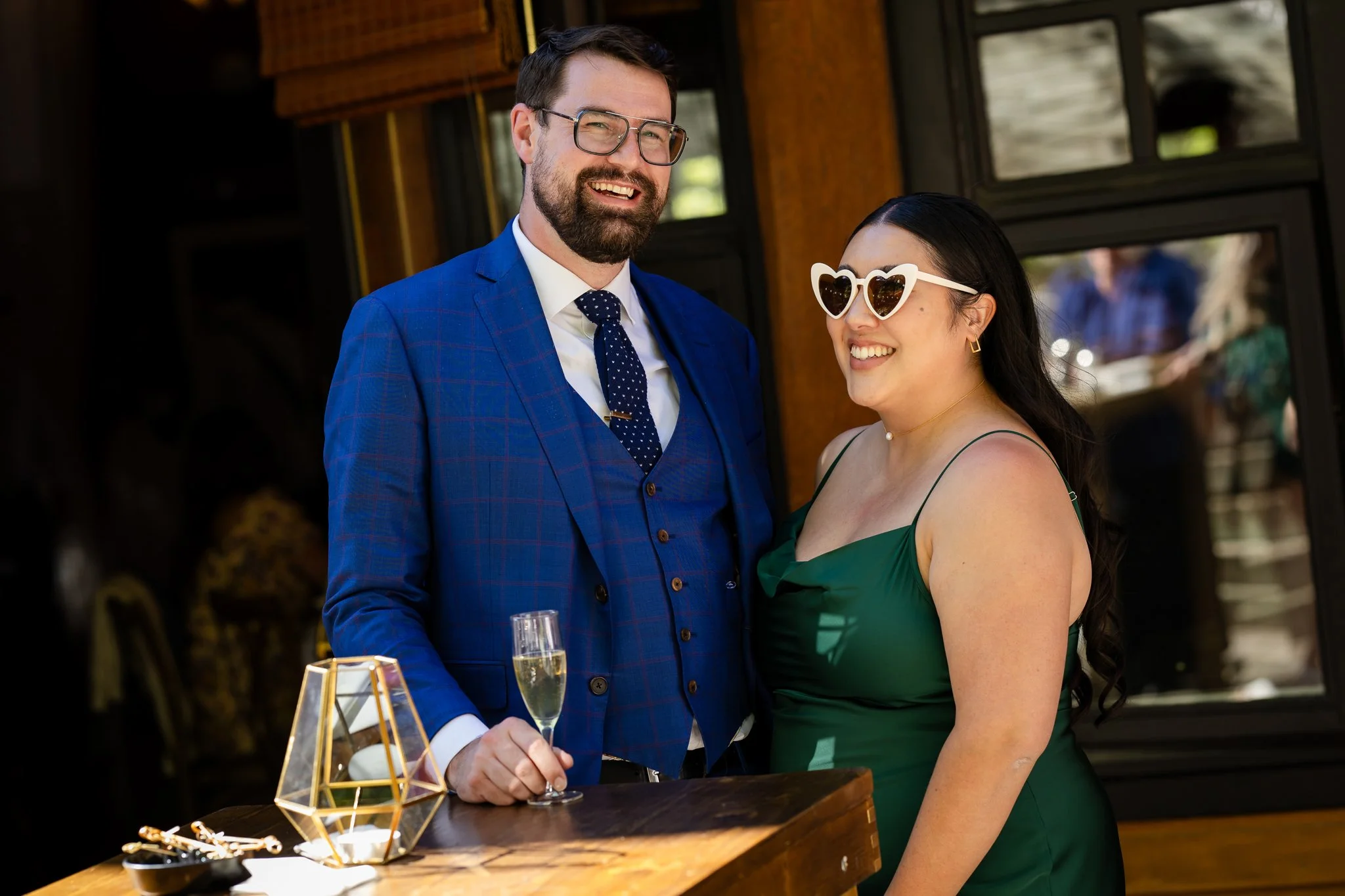 A man in a blue checkered suit, white shirt, dark dotted tie, and glasses, holding a glass of champagne, stands beside a woman in a green dress and heart-shaped sunglasses, both smiling at an indoor social event with dark wood accents and a window in