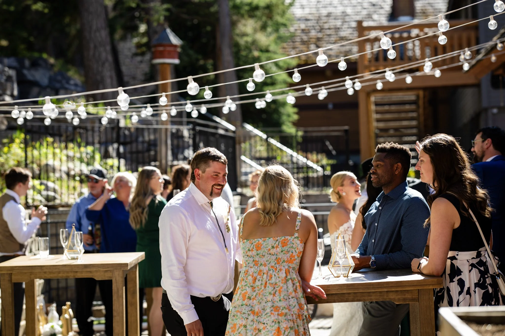 People socializing at an outdoor gathering, with string lights overhead and wooden tables.