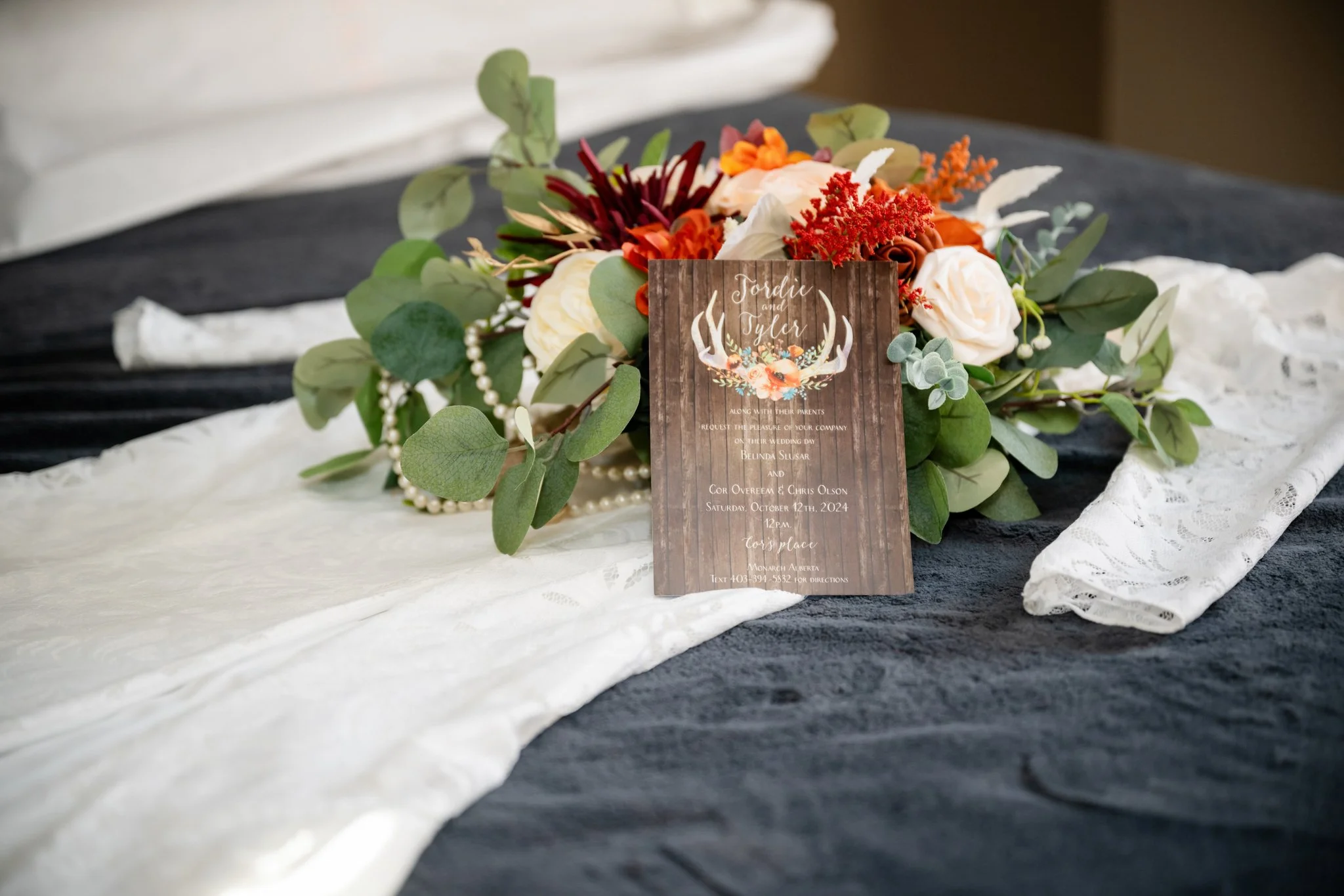Wedding invitation card placed on a floral centerpiece with white roses, green leaves, and orange flowers, on a black and white tablecloth.
